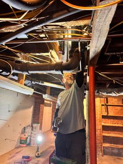 A person in a gray shirt stands in a cluttered basement, looking up while working on exposed overhead pipes.