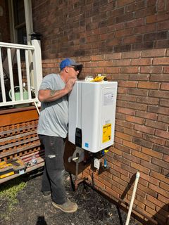 A technician installs a white tankless water heater on the exterior brick wall of a house.