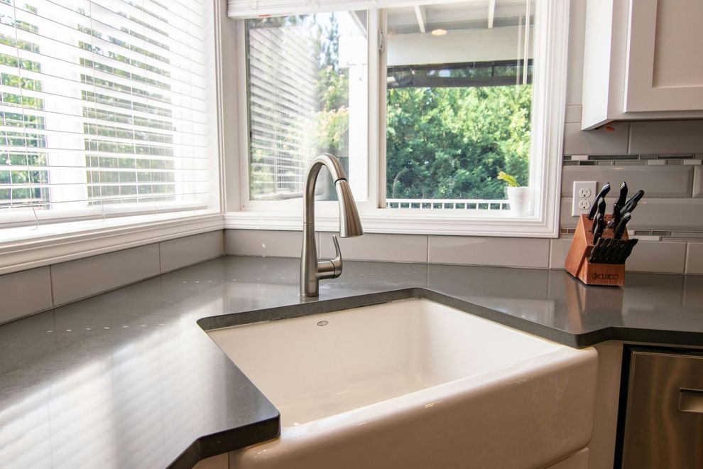 A white farmhouse kitchen sink with a chrome faucet set in a dark countertop under a window with white blinds.