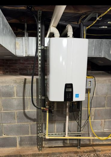 A white tankless water heater mounted on a metal strut frame against a cinder block basement wall with attached piping.