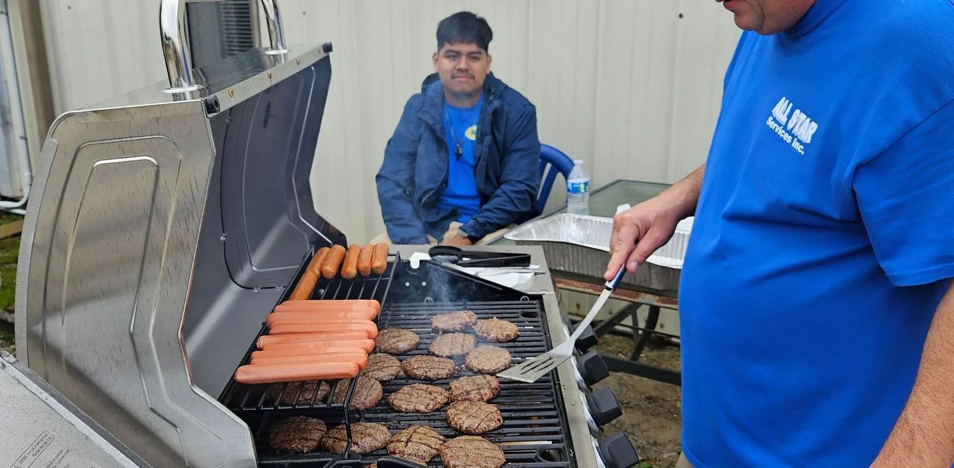 A man in a blue shirt is cooking hamburgers and hot dogs on a grill.