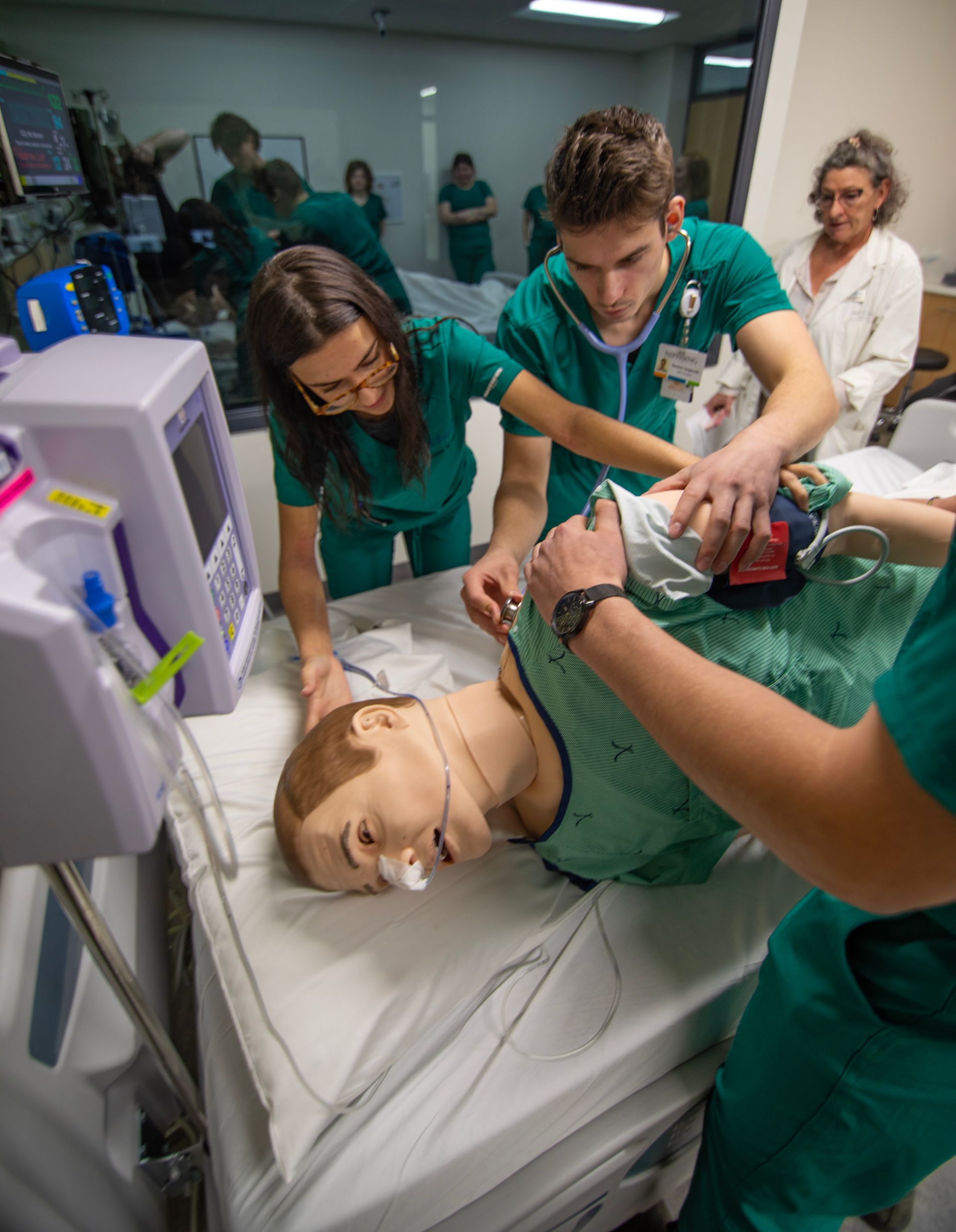 Nursing students practice on a medical dummy. They wear green scrubs, examining a patient in a hospital setting.