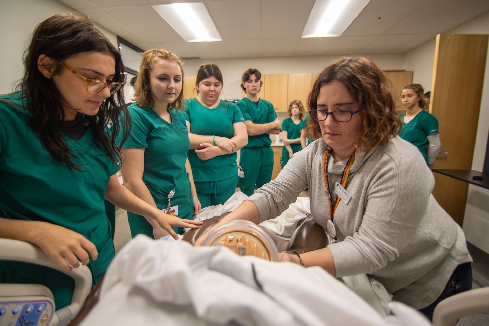 Nursing students in green scrubs watch an instructor demonstrate on a mannequin in a classroom.