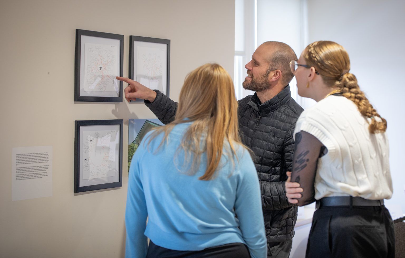 A man points at framed drawings on a wall, explaining them to two women in an office setting.
