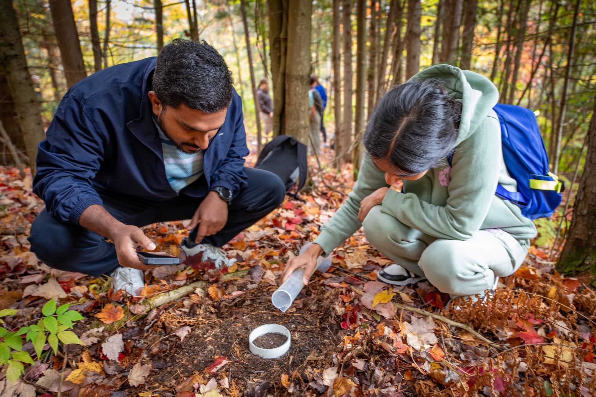 Two students in a forest collecting soil samples: one using a small container, the other pouring soil into a tube.