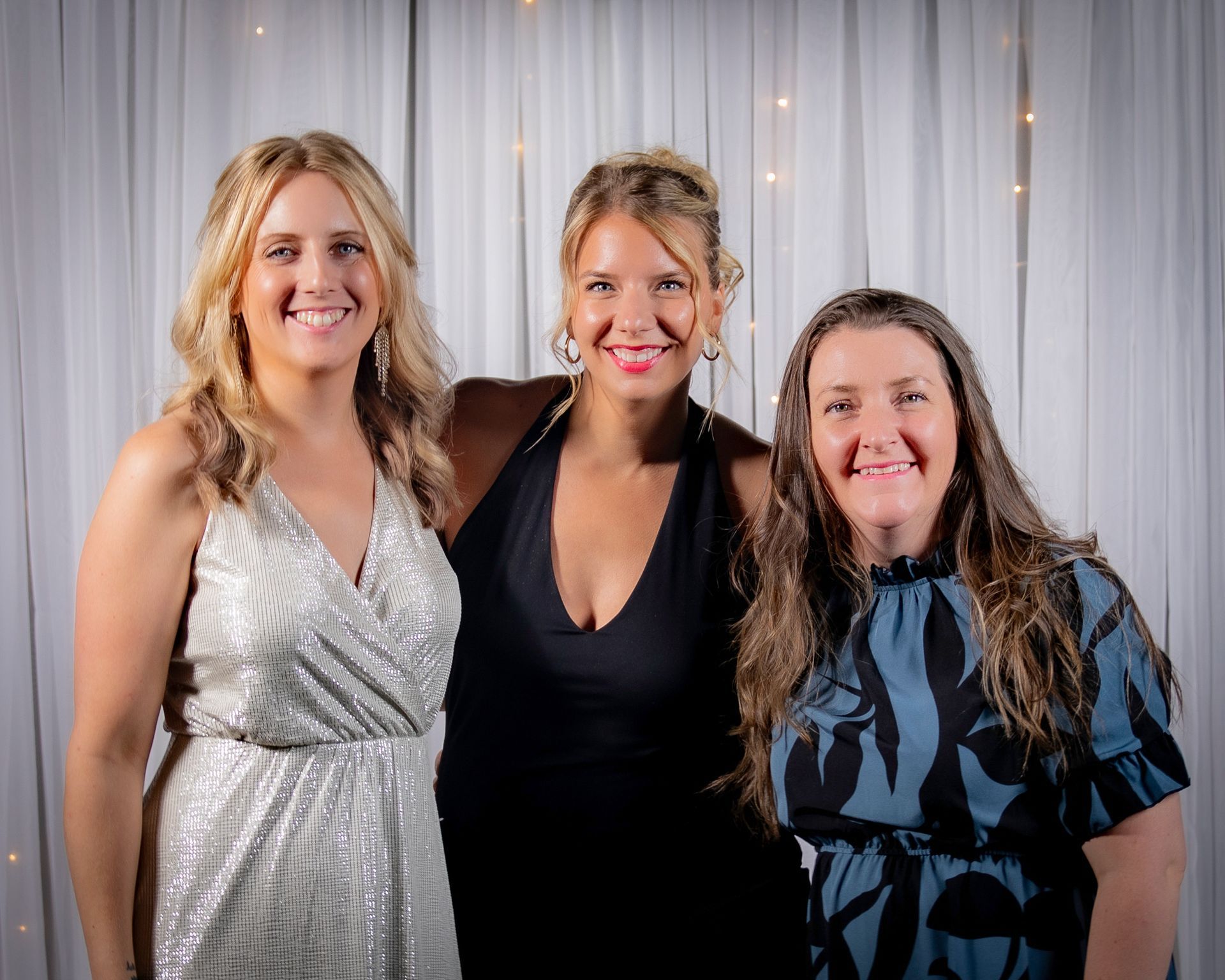 Three women smiling in formal attire in front of a draped white backdrop.