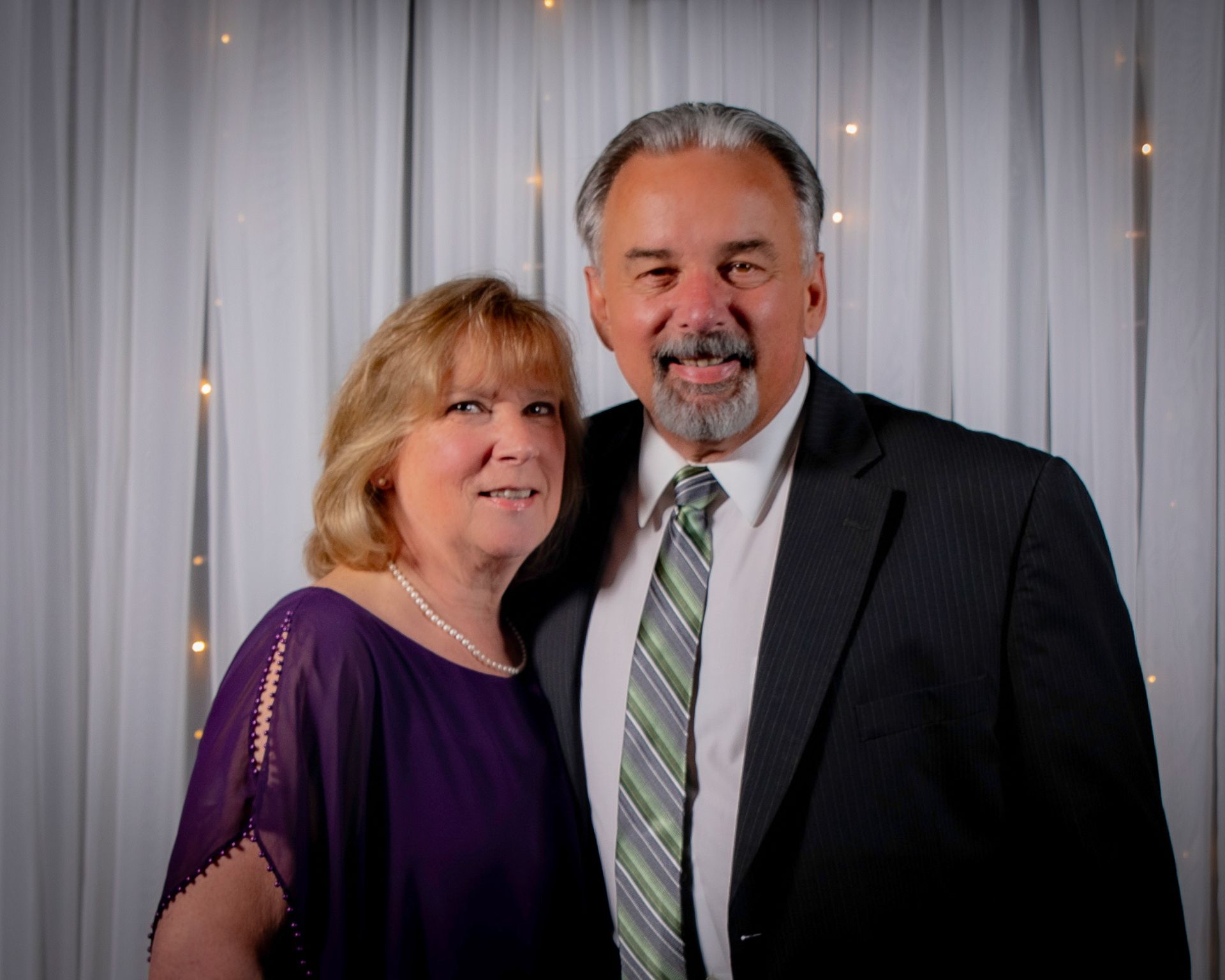 Couple, woman in purple dress and man in suit, smiling in front of white curtain with lights.