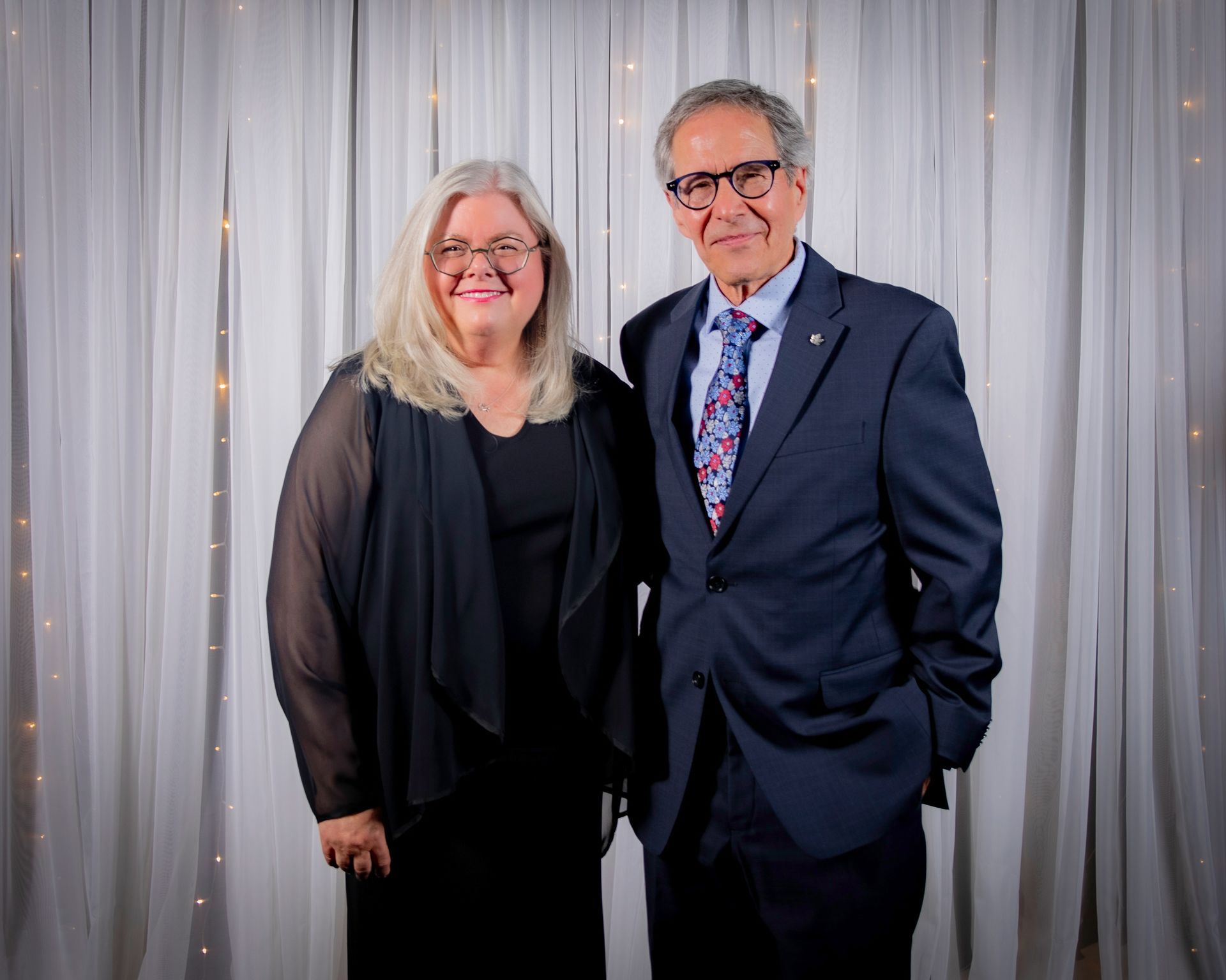 Woman with gray hair and man in suit pose in front of a white curtain.