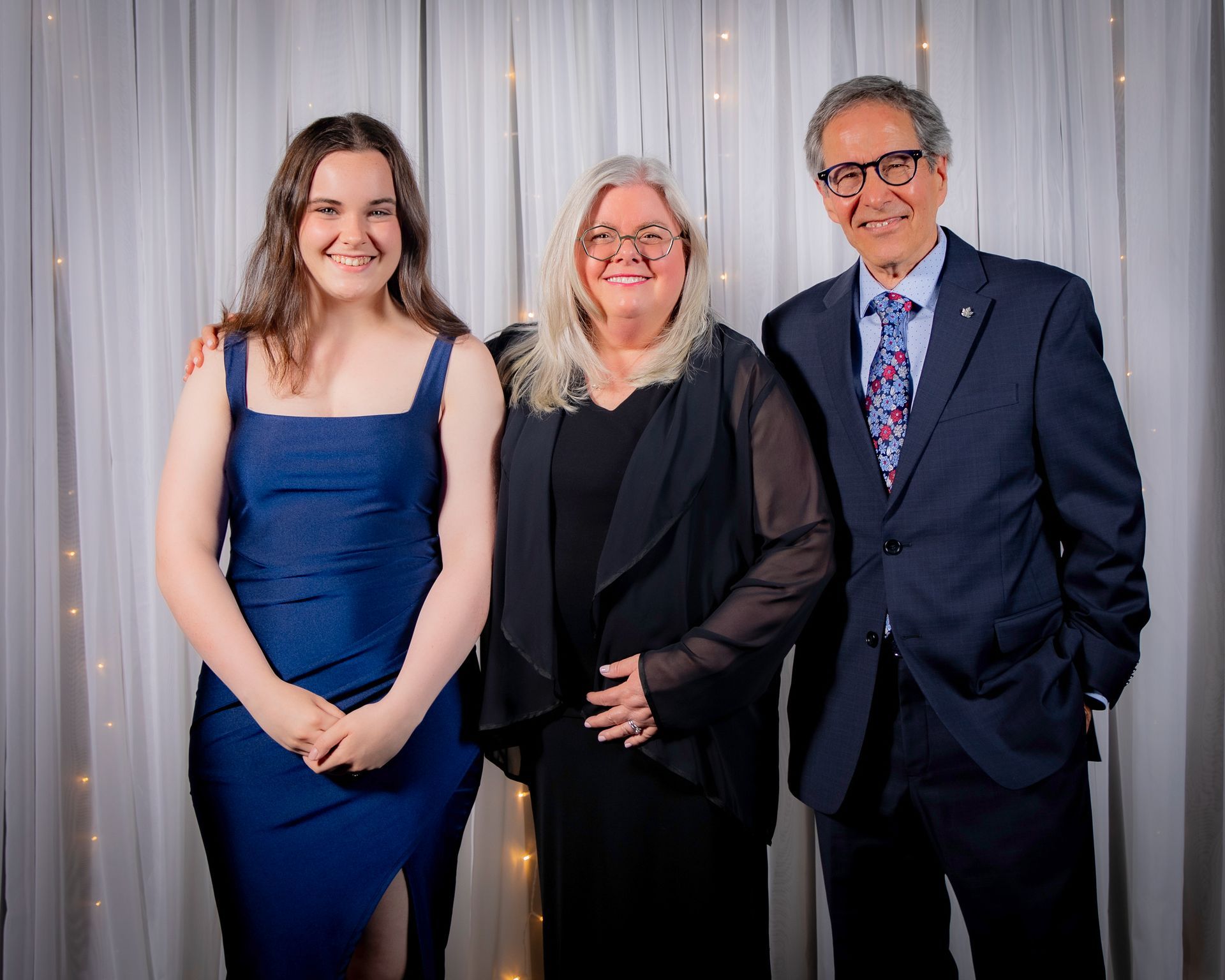 Three people smiling, posing in front of a white curtain. Woman in blue dress, woman in black, man in suit.