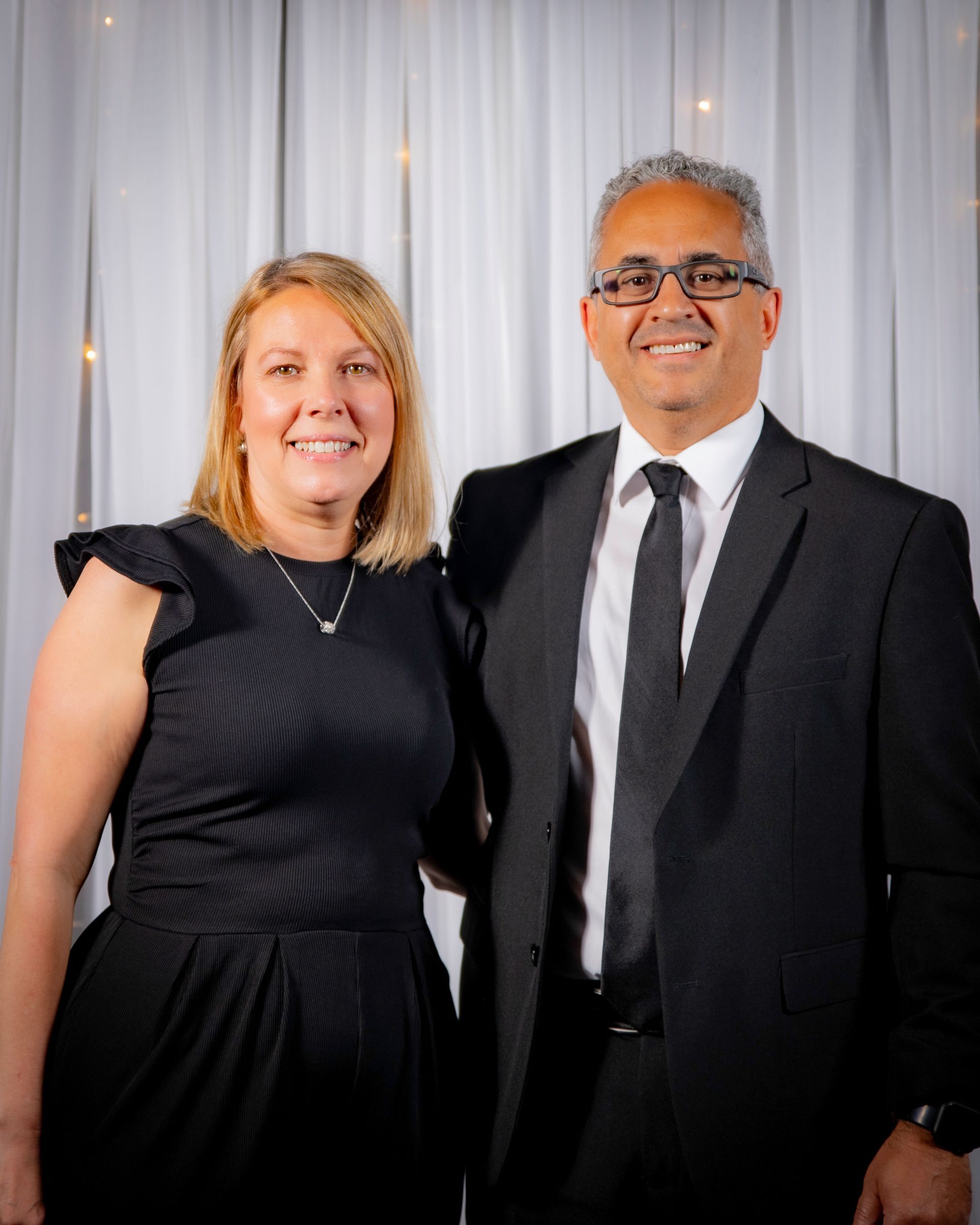 Woman and man smiling, posing together; the woman in a black dress, the man in a suit. White backdrop.