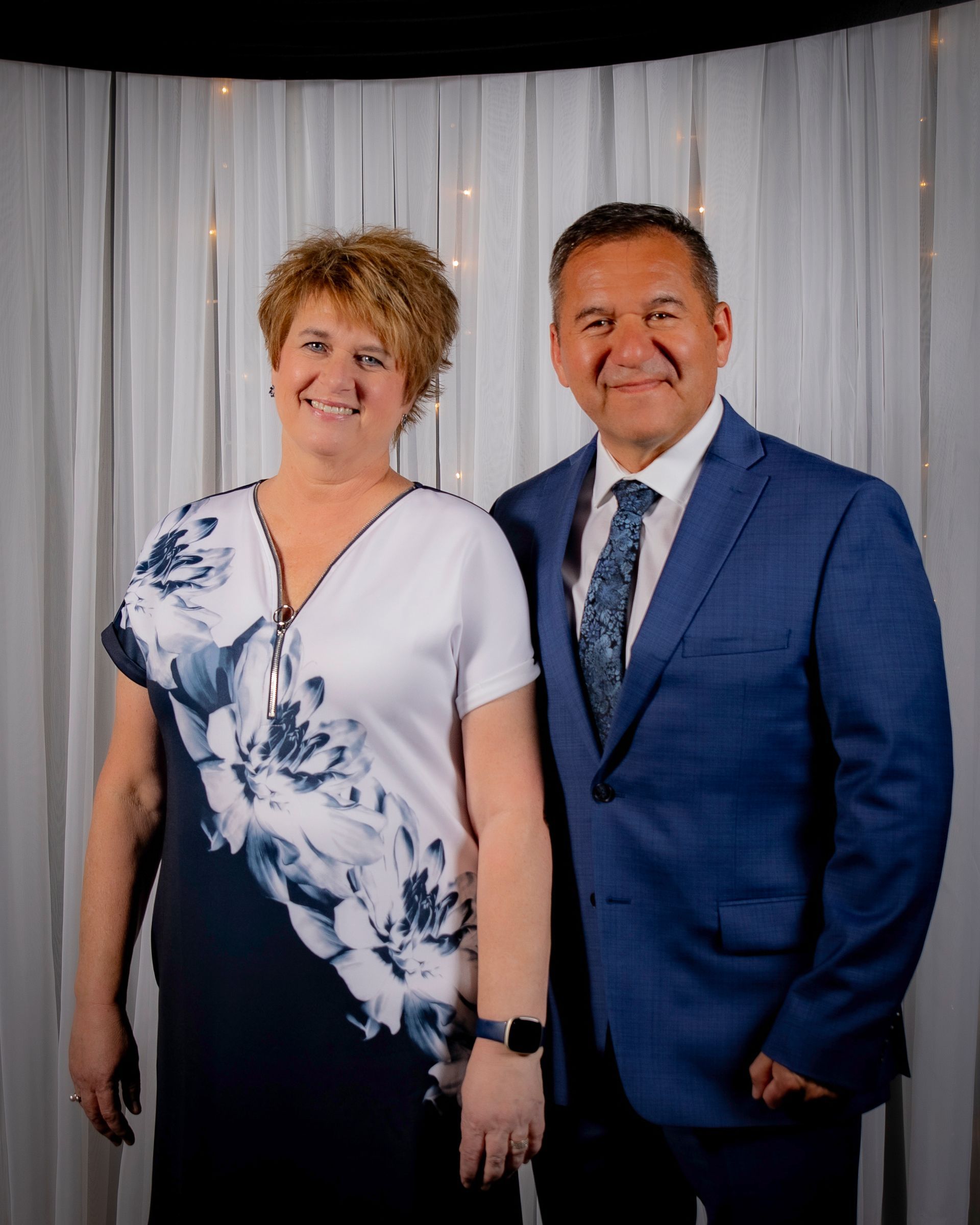 Couple smiling, posing in front of a white curtain backdrop. Woman in floral dress, man in blue suit.