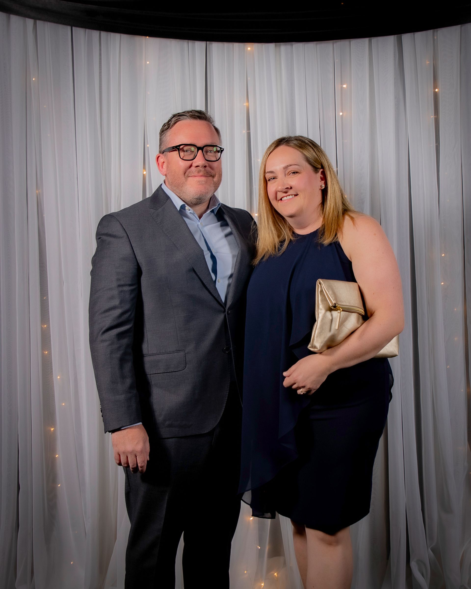 Man and woman in formal attire stand in front of a white curtain, man wears glasses and a suit, woman in navy dress.