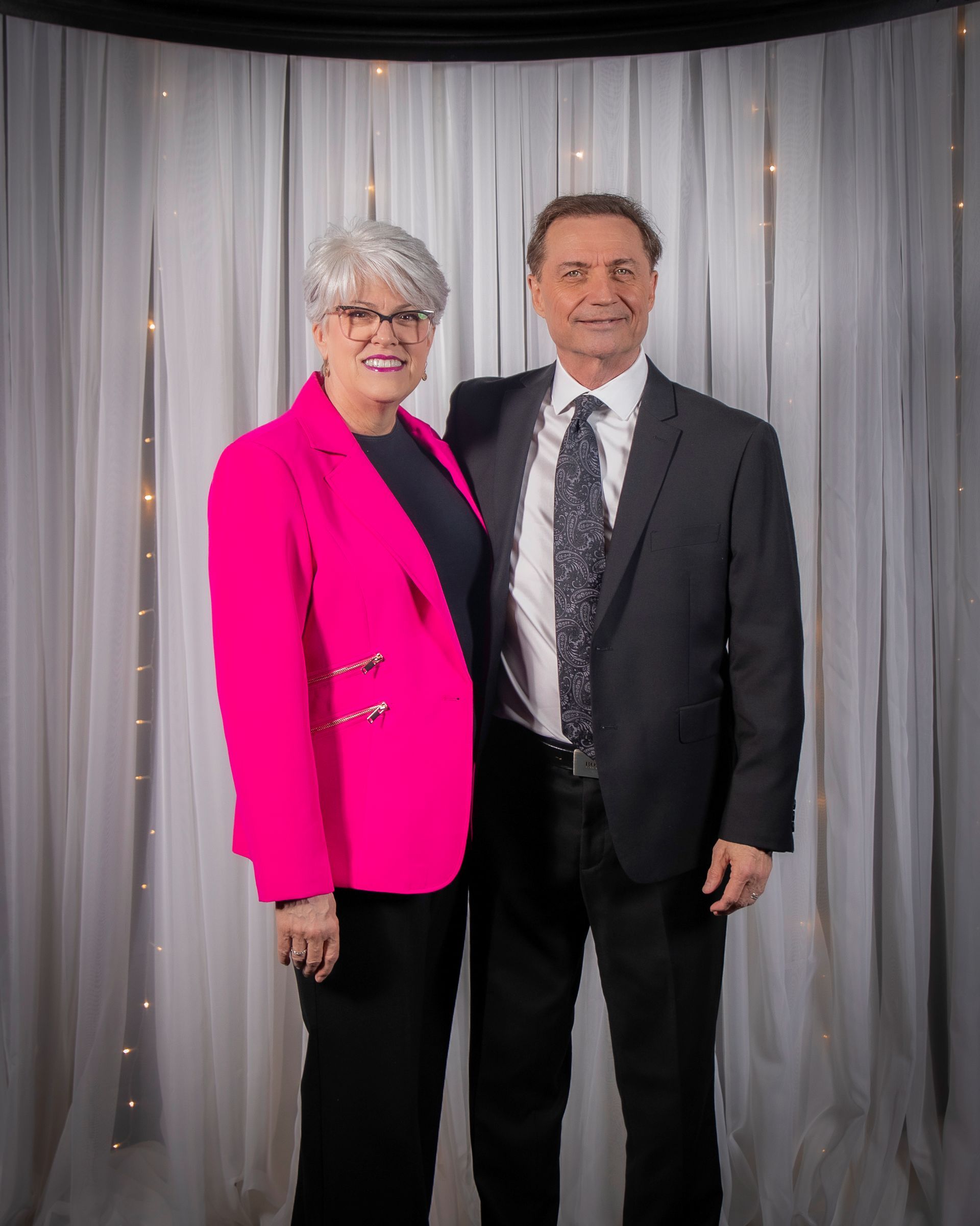 Couple in formal attire standing together in front of a curtain backdrop, smiling. The woman wears a pink blazer.