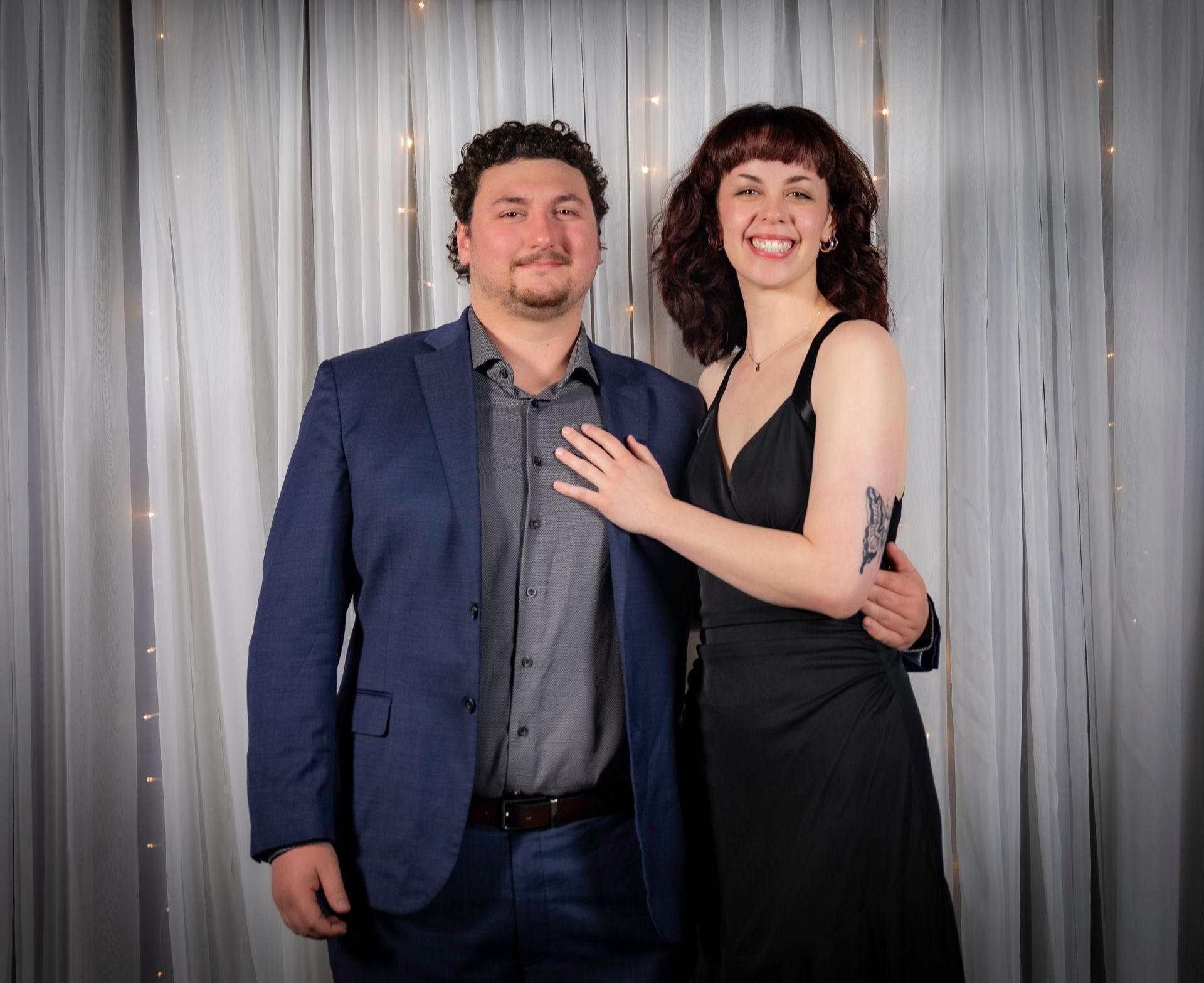 Man in blue suit and woman in black dress pose smiling in front of a white curtain backdrop.