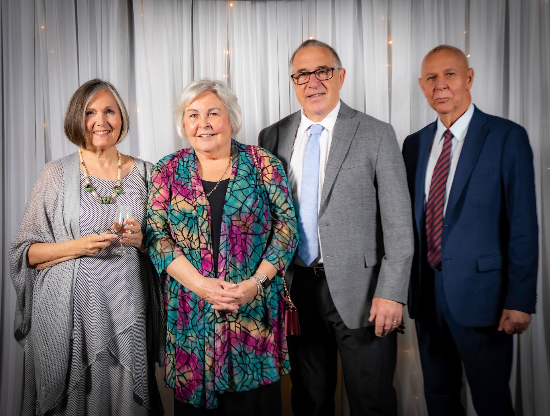Four people smiling, posing together in front of a white curtain with lights.