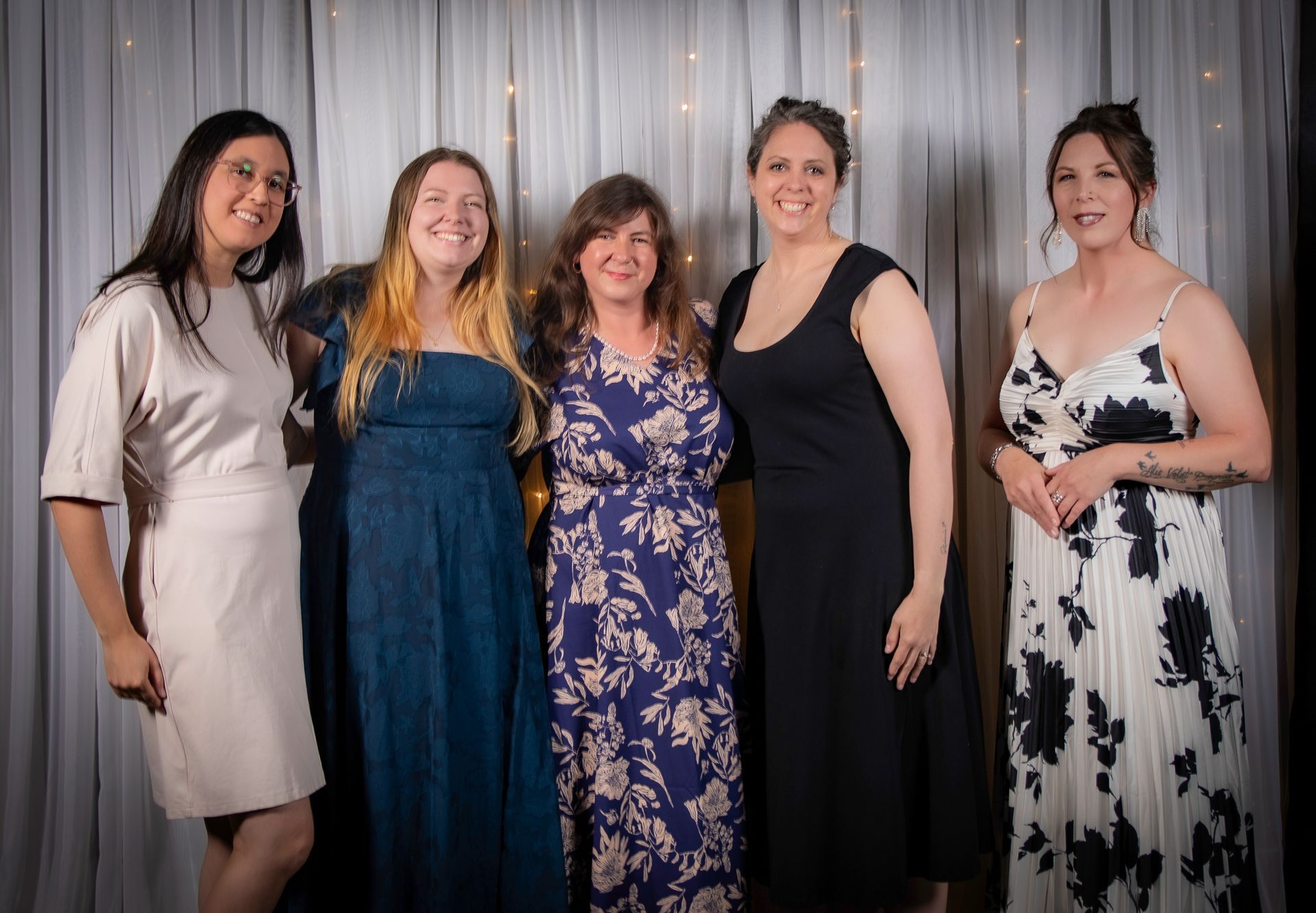 Five women smiling, posing for a photo in front of a white curtain backdrop.