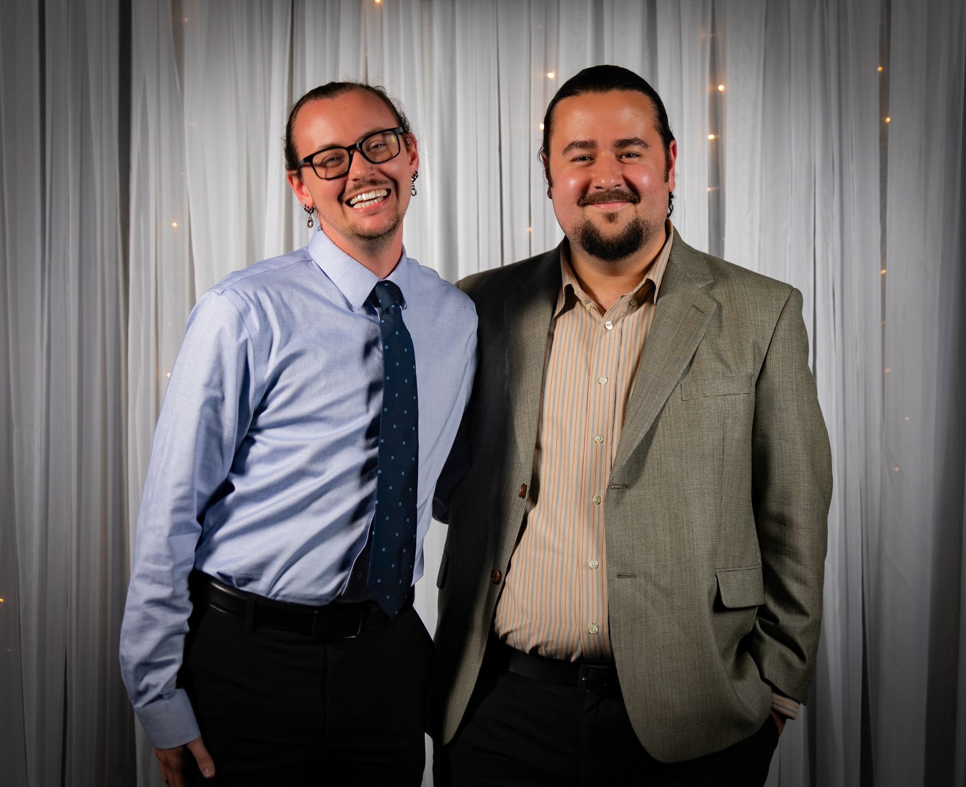 Two men smiling, standing together in front of a white curtain with lights.