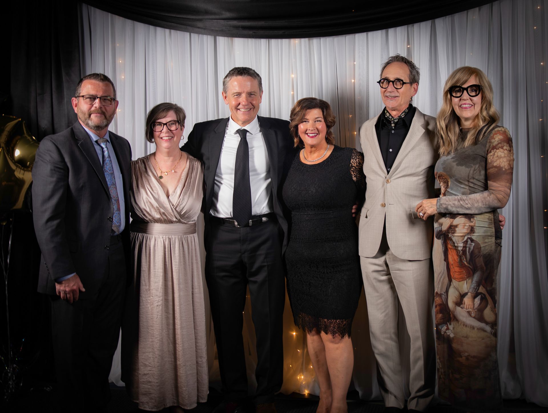 Group of six people posing in formal wear, smiling, in front of a curtain backdrop.