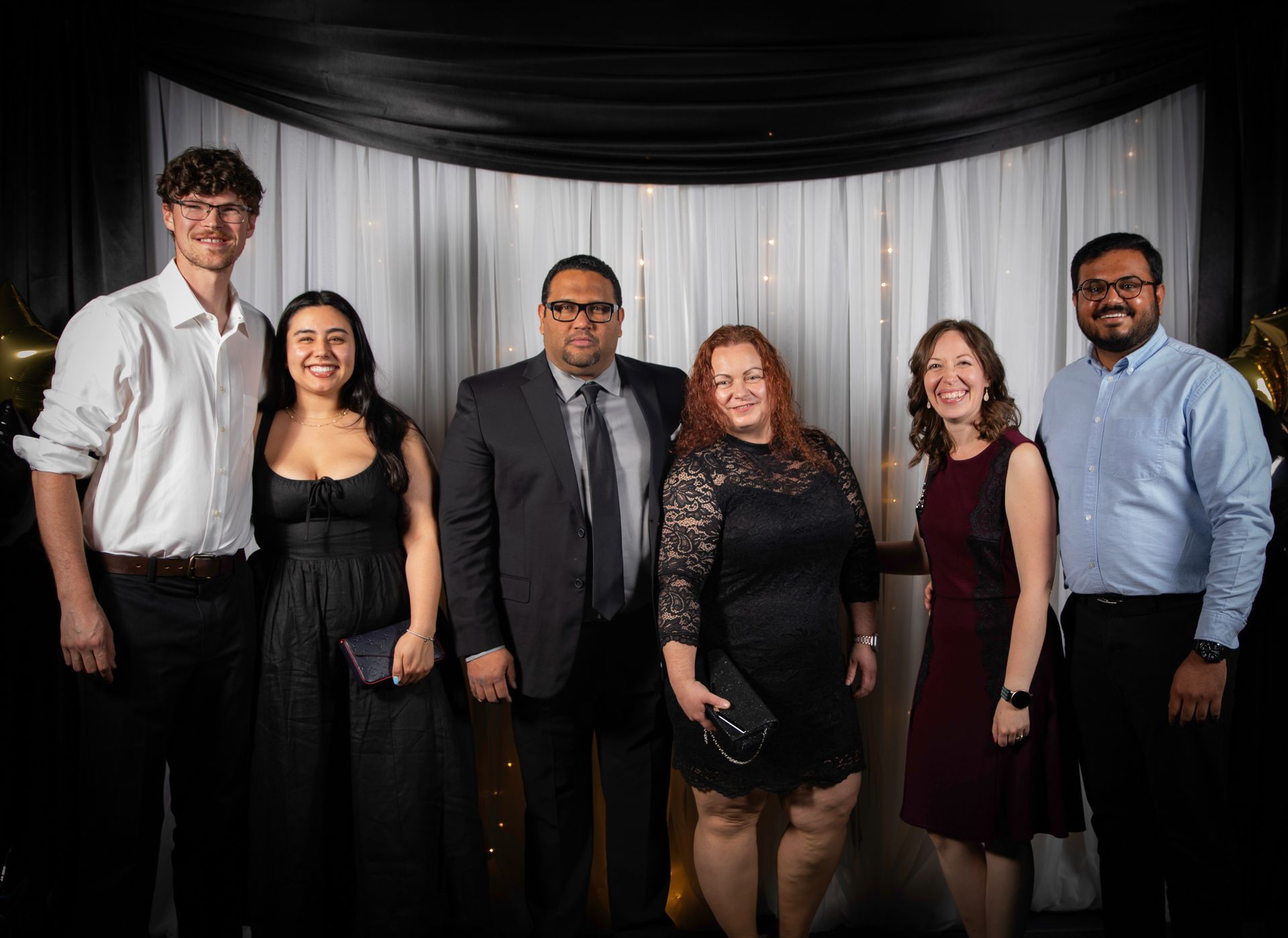 Group of six people posing for a photo against a backdrop of draped fabric, smiling, formal attire.
