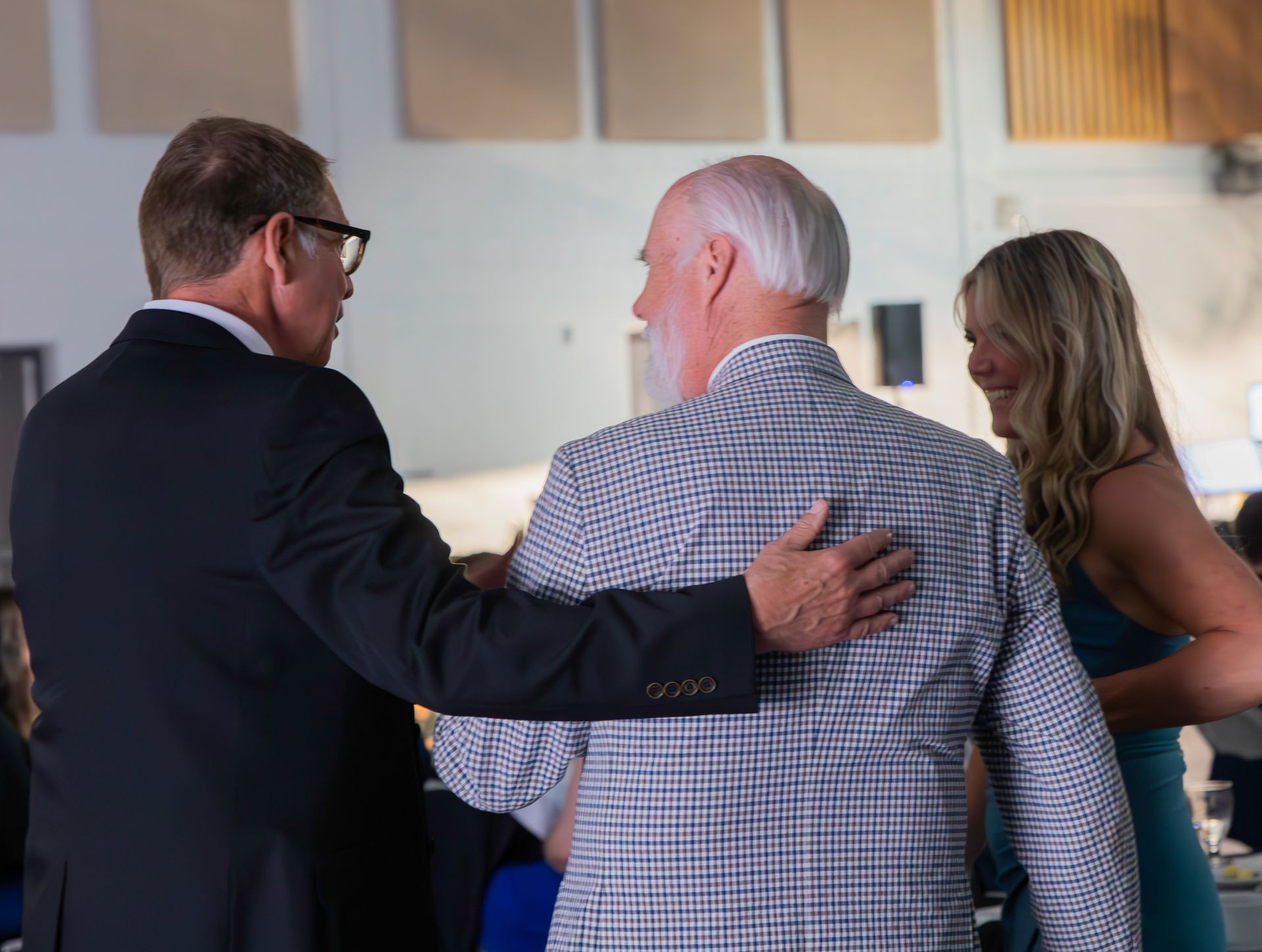Three people in a room; two men in suits and a woman in a blue dress. Man in suit has hand on the other man’s back.