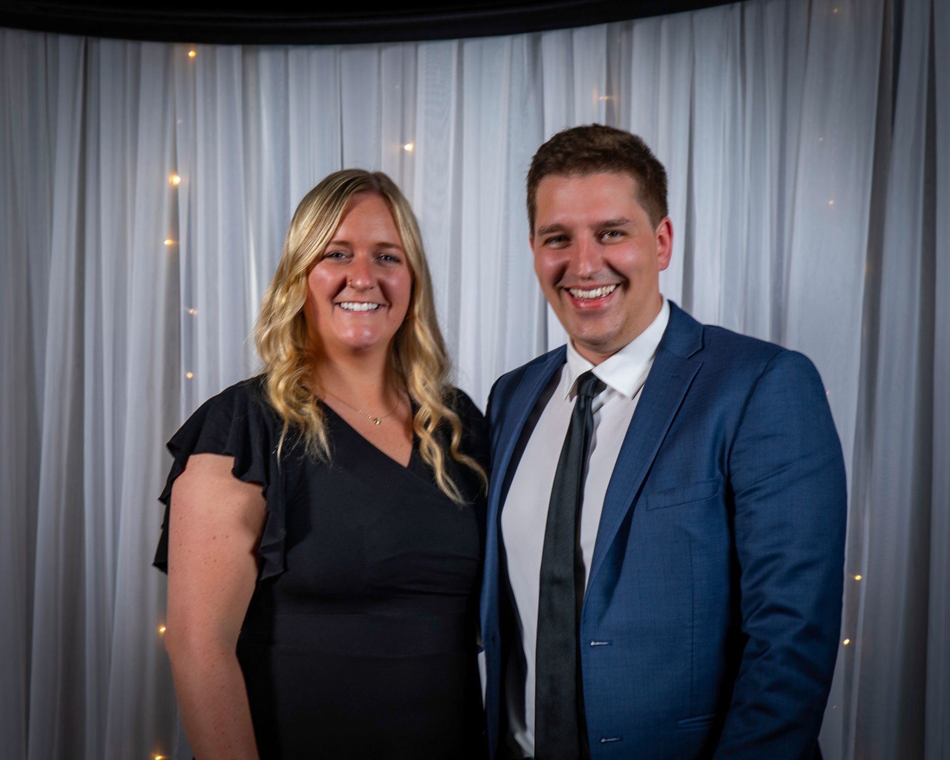 Woman and man smiling, posing together in front of a white curtain with lights. Man is wearing a suit.