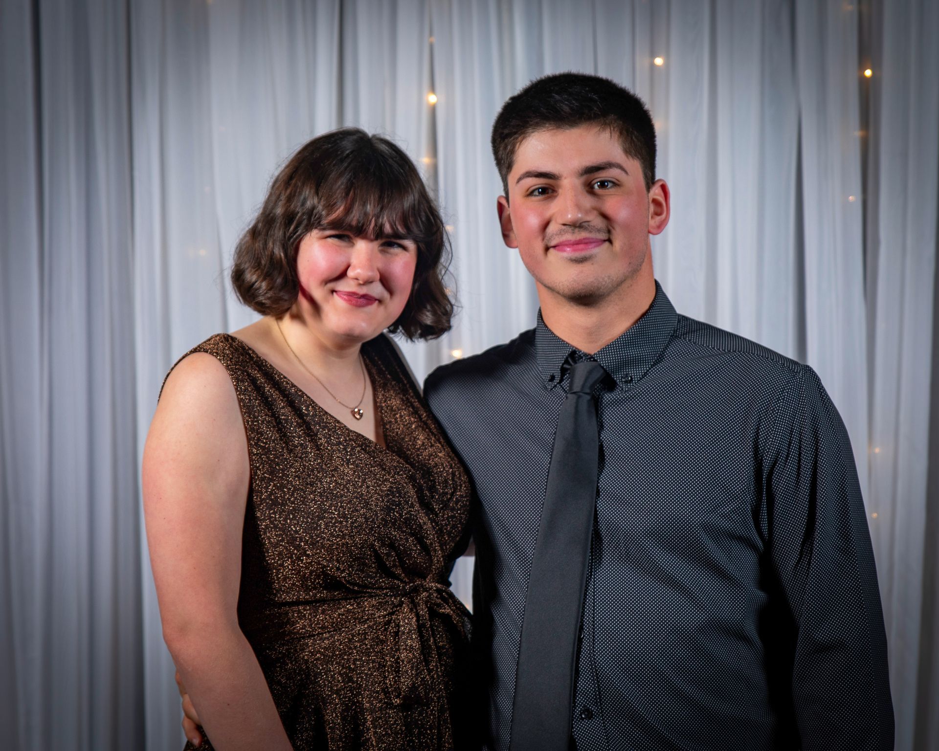 Young couple smiles in front of a white curtain backdrop. Woman in brown dress, man in patterned shirt with black tie.