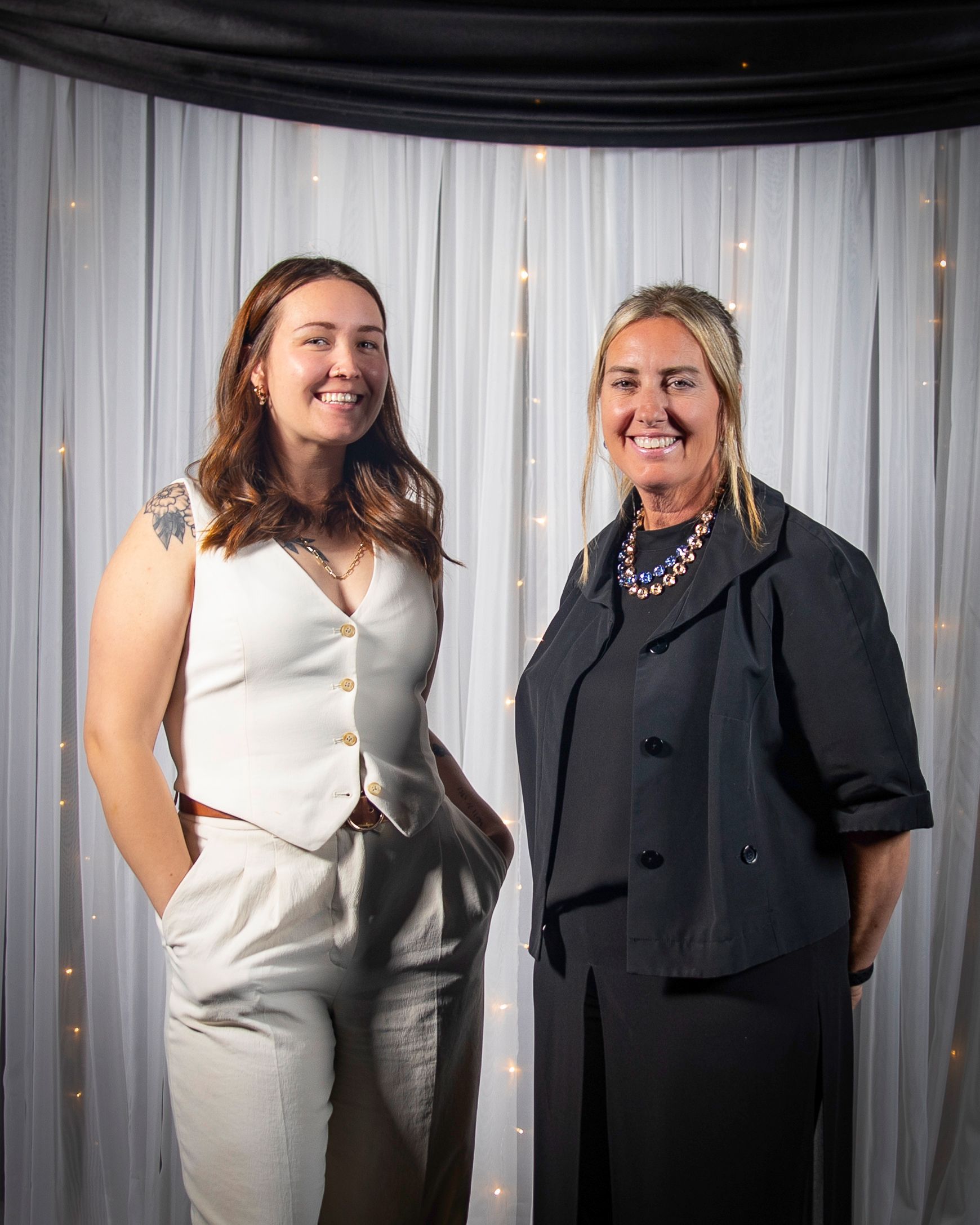 Two smiling women posing in front of a white curtain. 
