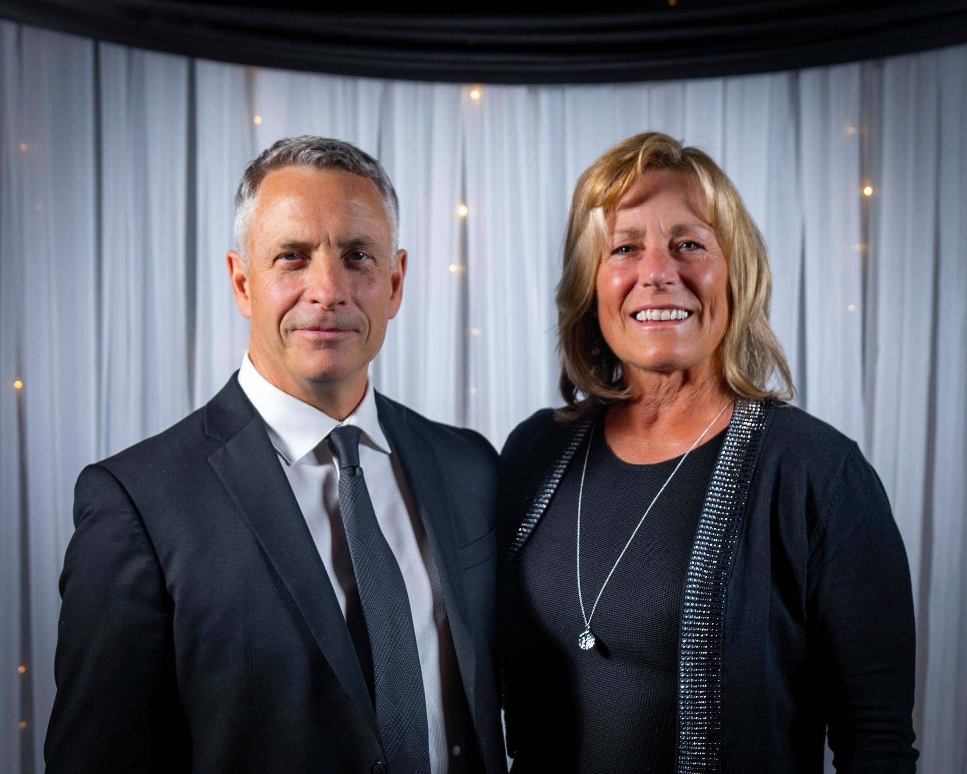 Man and woman in formal attire, posing and smiling in front of a lit backdrop.