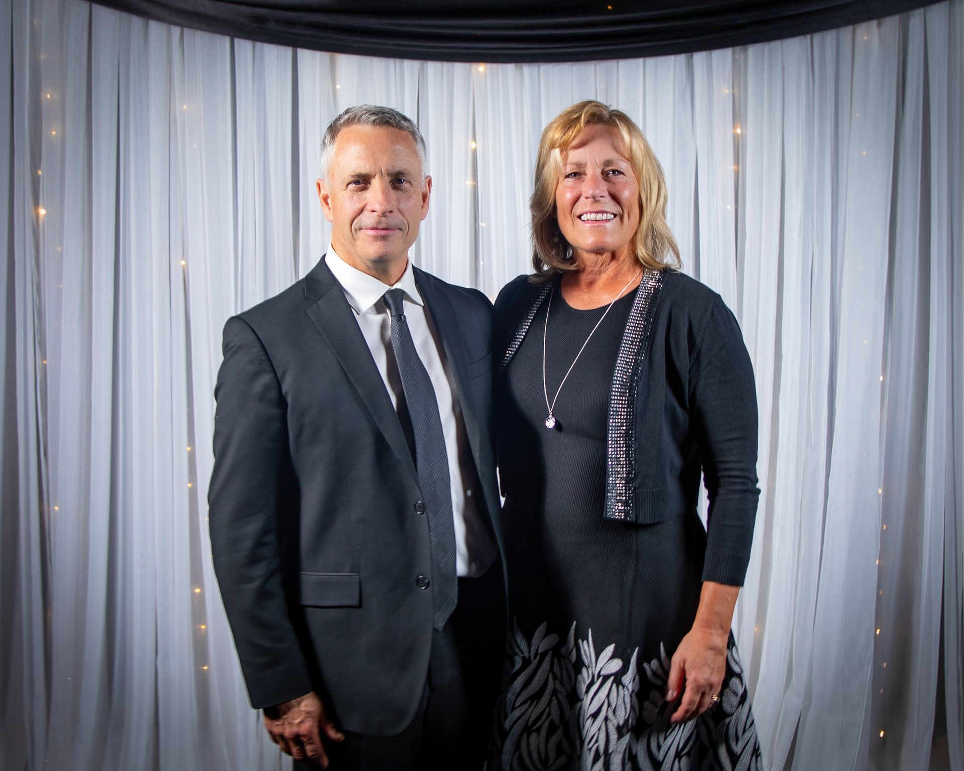 Man and woman pose formally, in front of a draped backdrop, smiling. Man wears a suit, woman a dress.