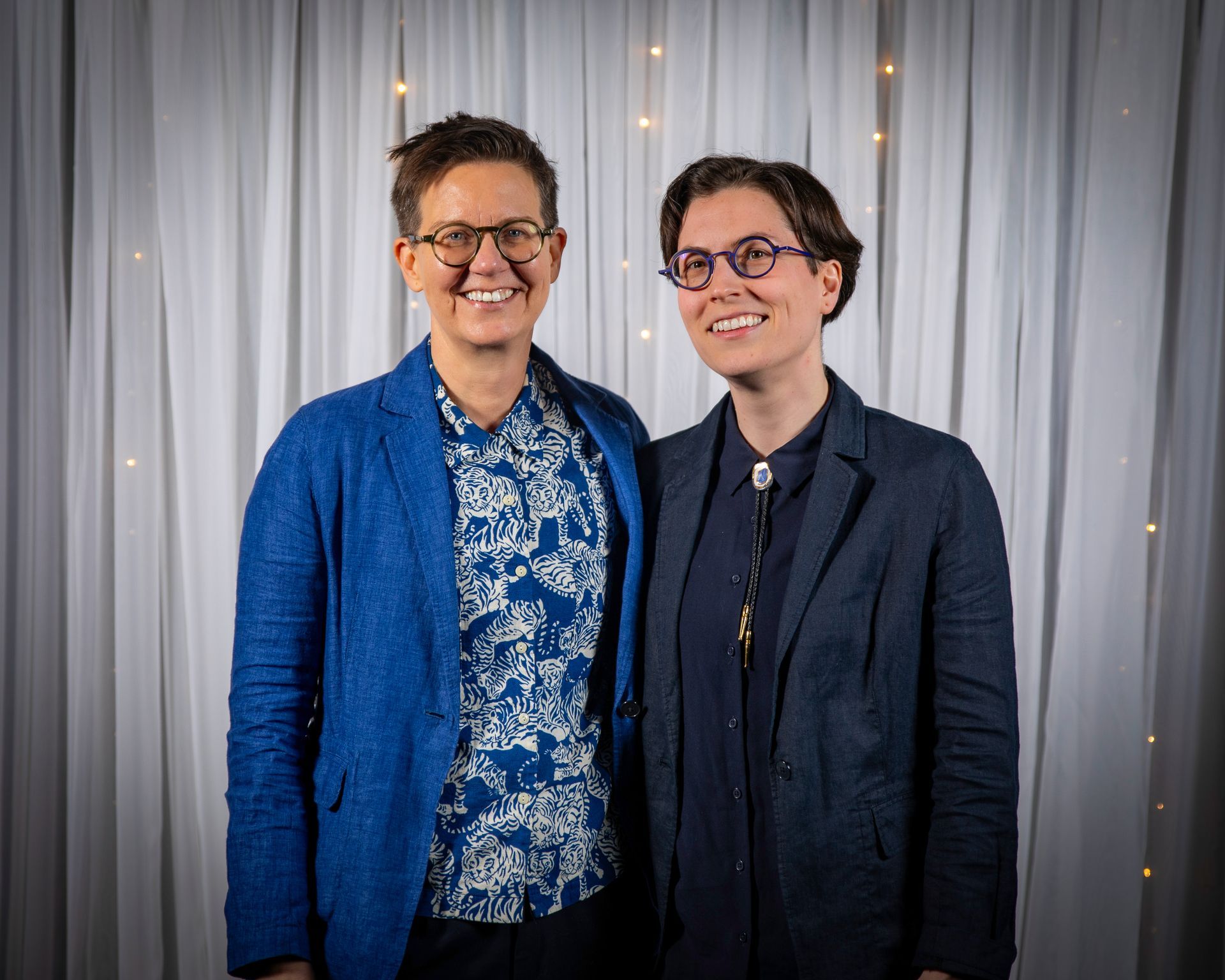 Two people, smiling, standing in front of a white curtain. They both wear glasses and blazers, one blue, the other navy.