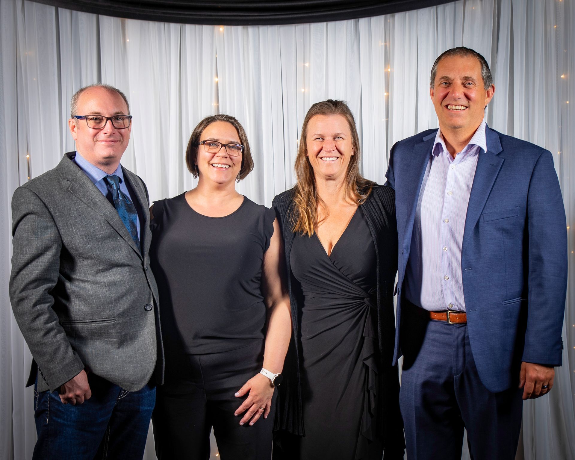 Four people smiling for a photo in front of a white curtain. Men in blazers, women in dark tops.
