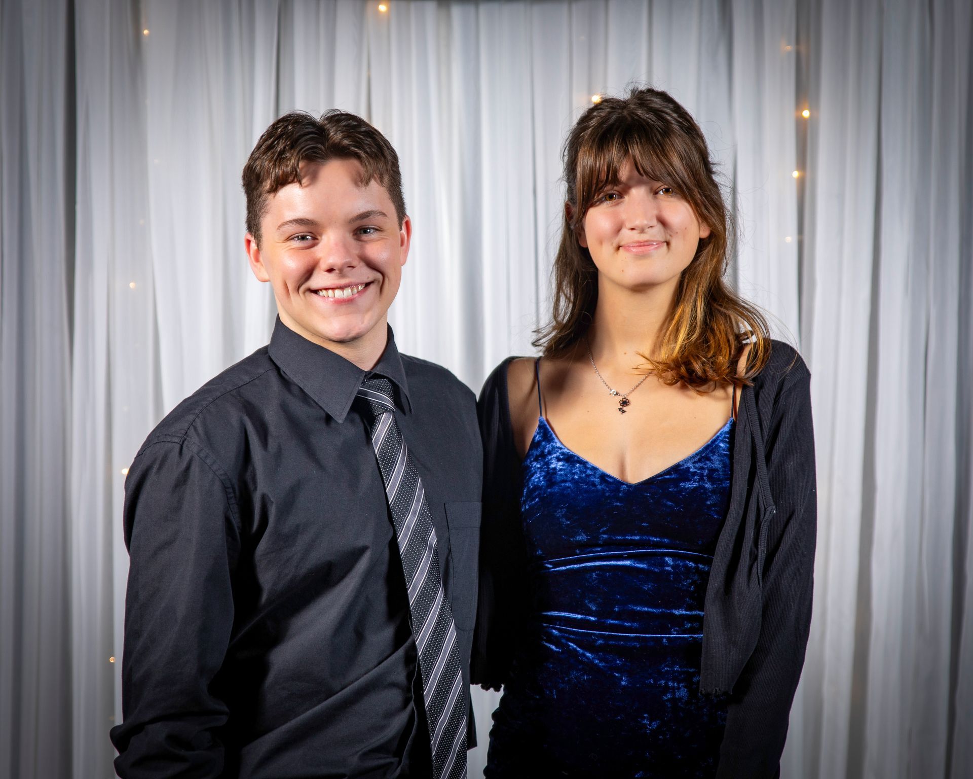 A young man and woman smiling at a formal event. The woman wears a blue dress, the man a black shirt and tie.