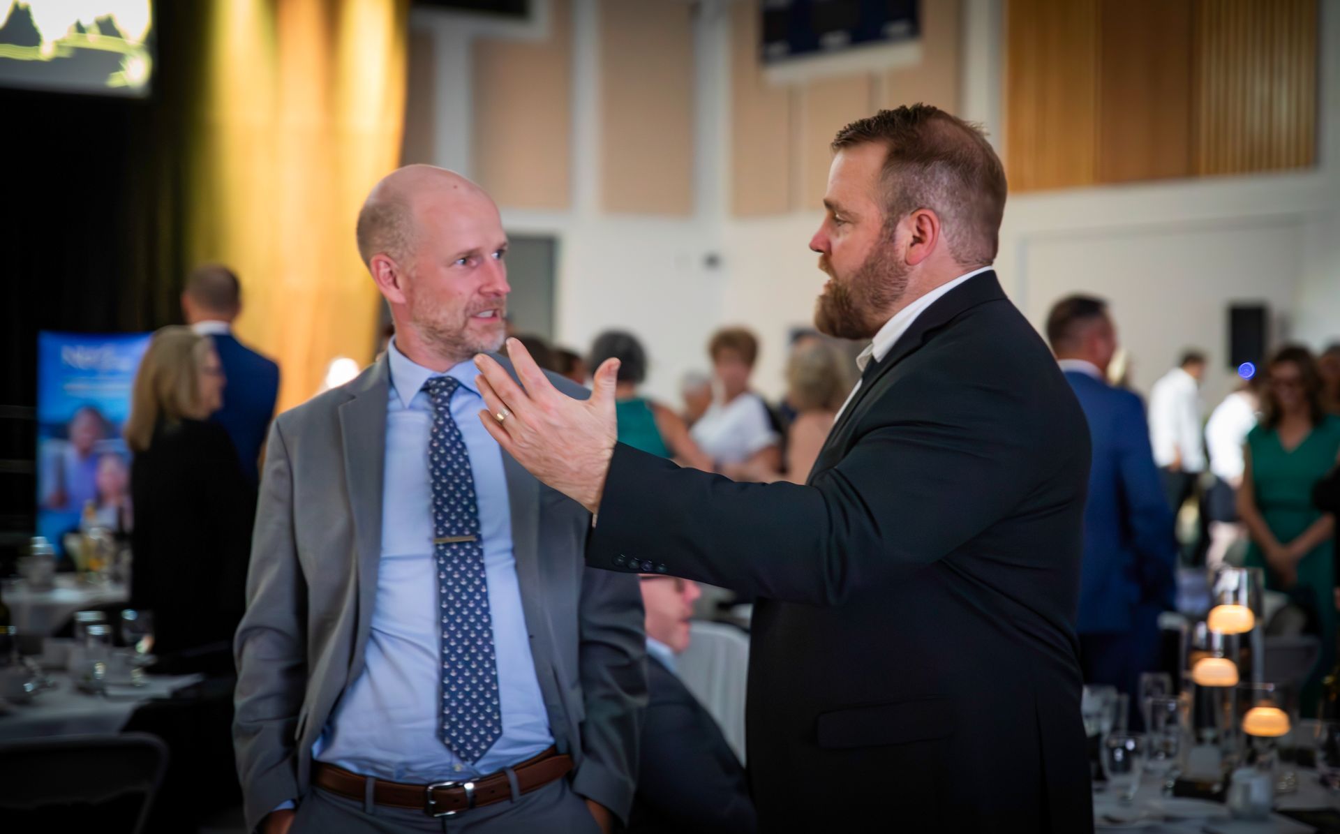 Two men in suits conversing at a formal event. One gestures with hand, others in the background.