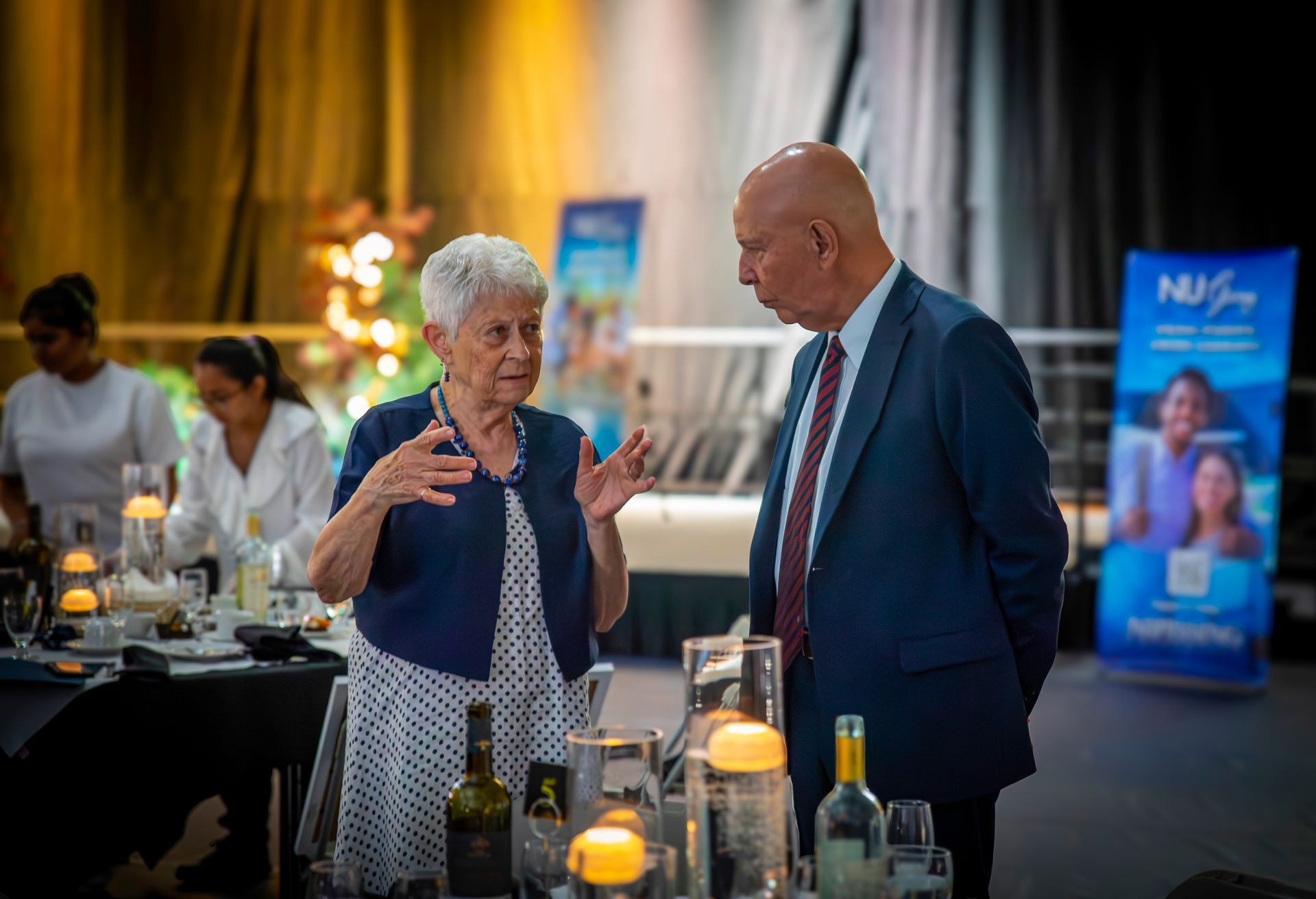 Two people in conversation at a formal event, tables set for dining, staff in background, blue banners.
