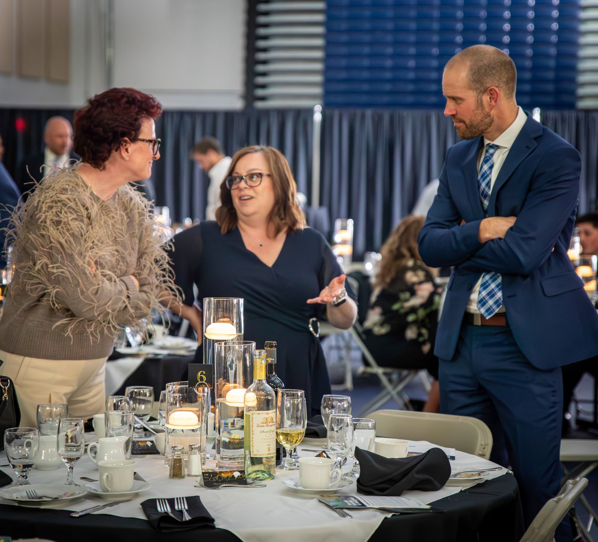 Three people conversing at a formal event. Woman in beige sweater gestures, other woman speaks, man in suit listens.