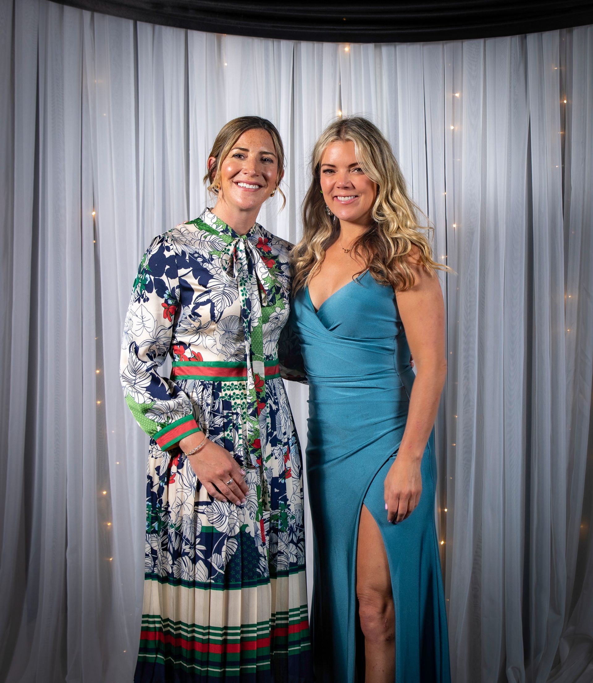 Two women at an event pose in front of a curtain. One wears a floral dress, the other a blue gown.