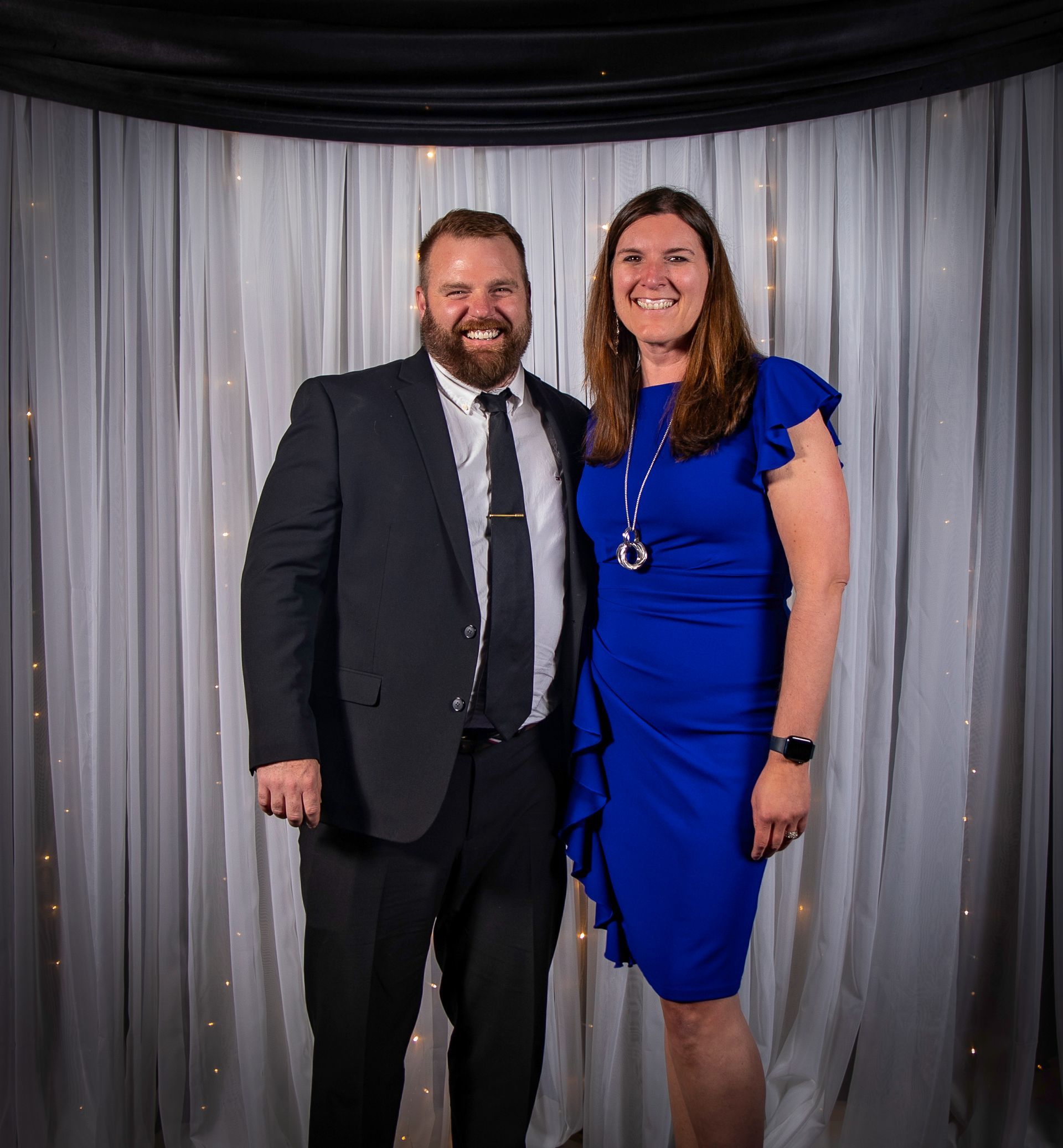 Man and woman smiling, posing together in front of white curtains, man in suit, woman in blue dress.