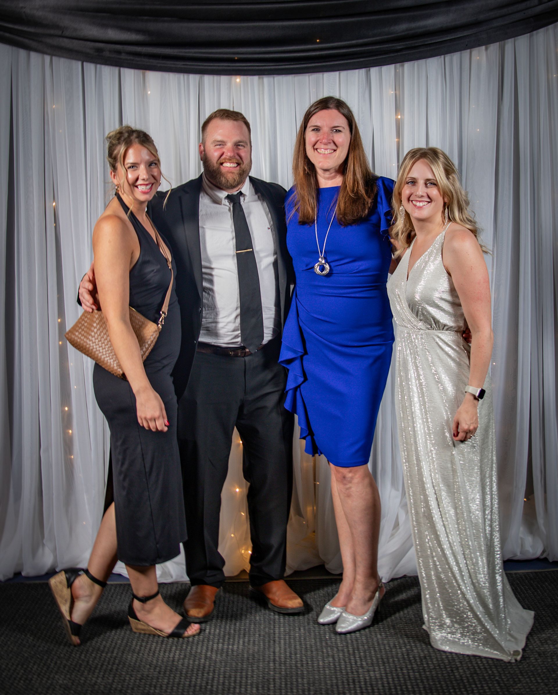Four people smiling, posing for a photo. Two women wear gowns, one blue, one silver. Man in suit. Neutral backdrop.