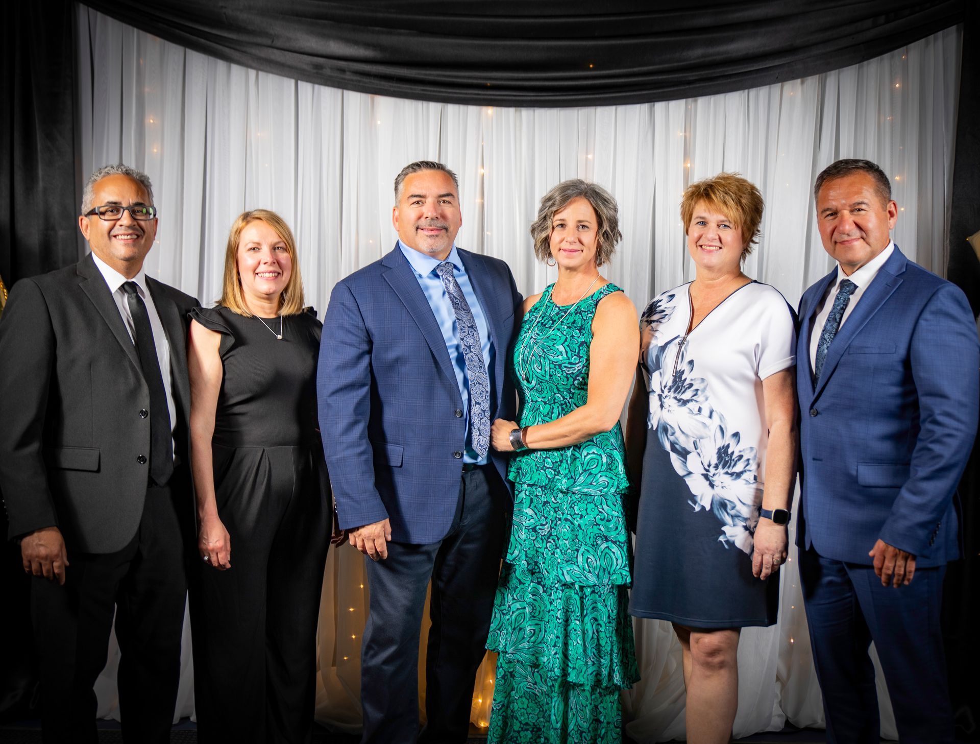 Group of six people, formal attire, posing for photo, smiling in front of draped backdrop.