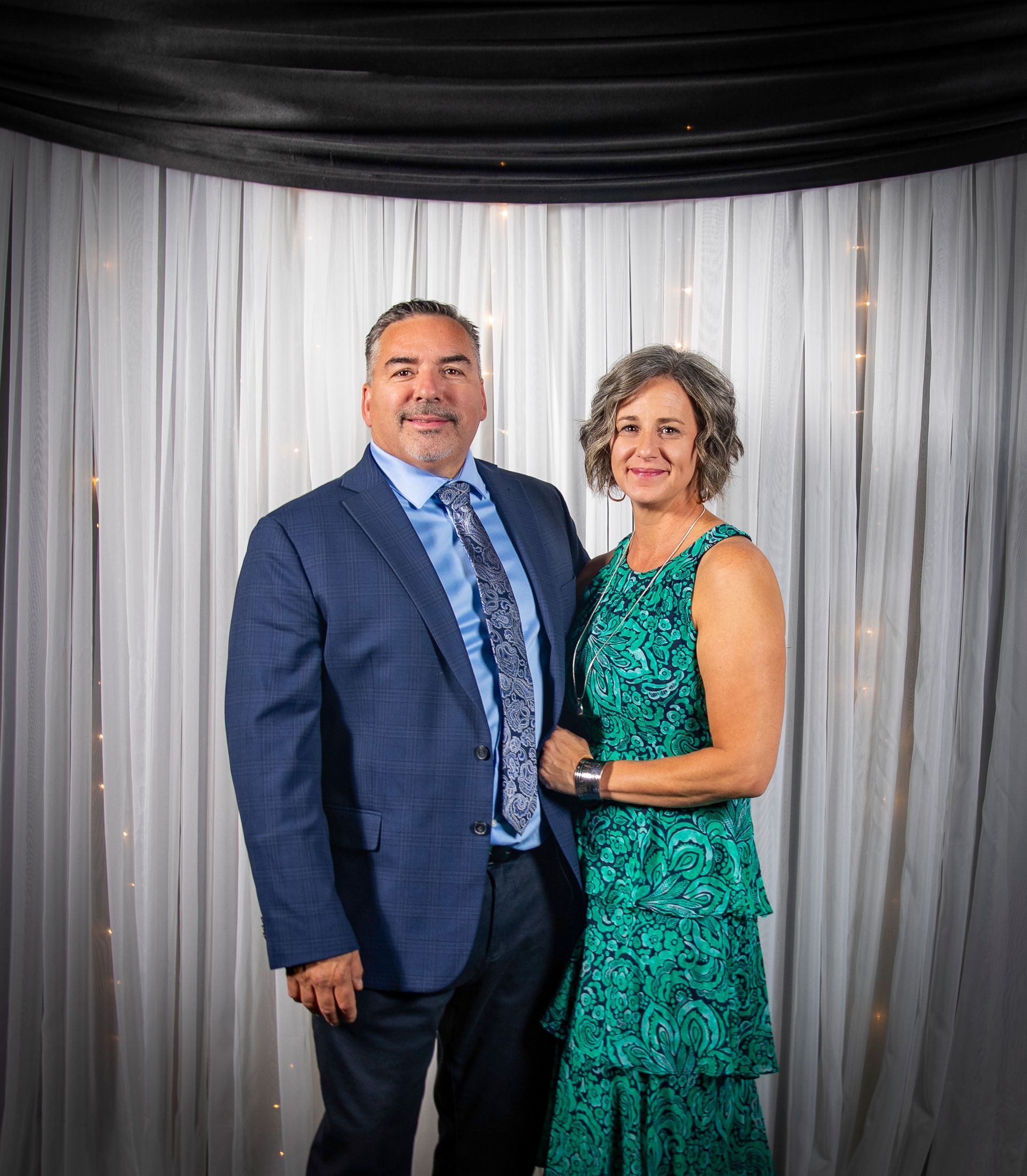 Couple posing for a formal photo. Man in blue suit, woman in green dress; background with curtains.