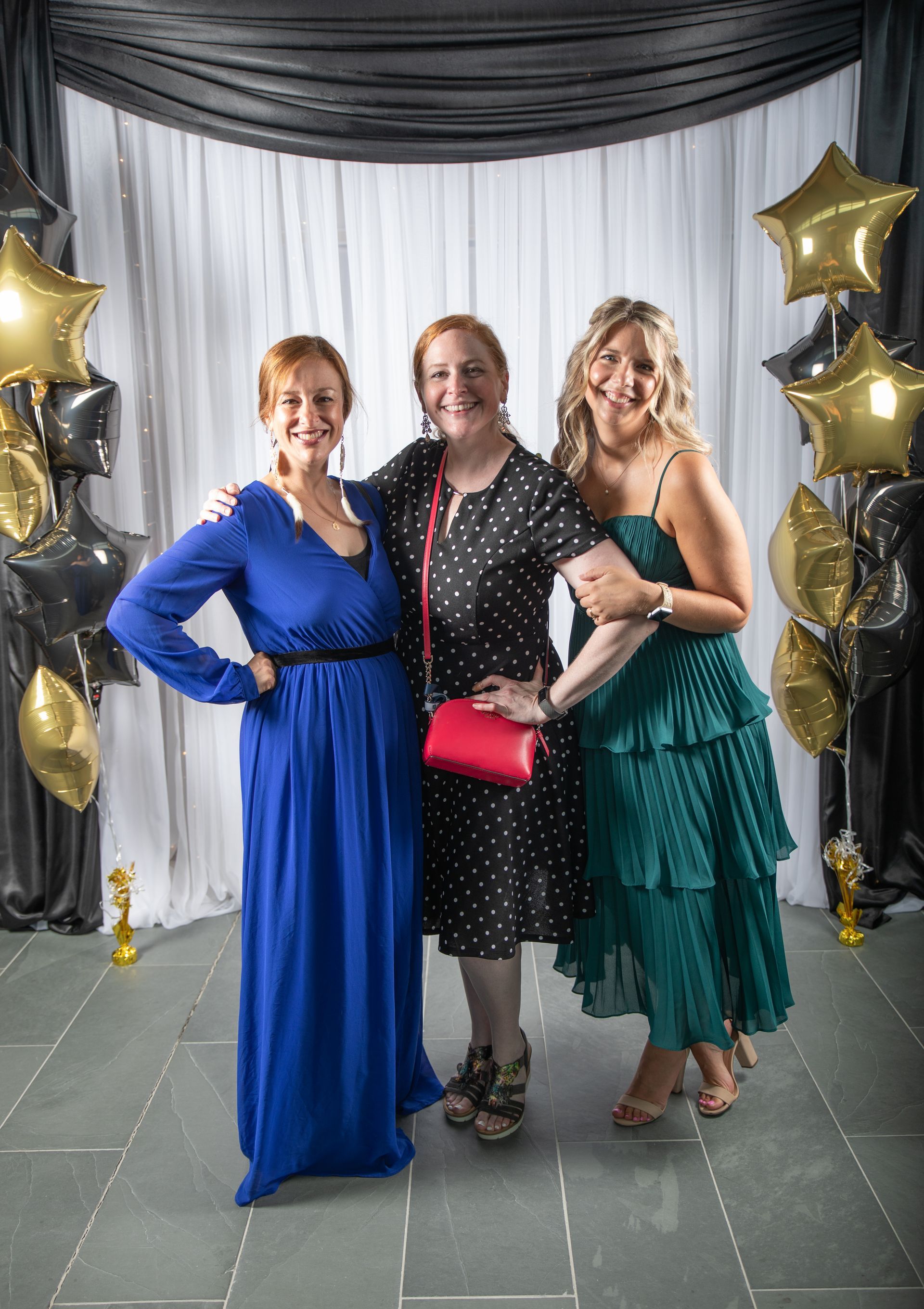 Three women in formal attire pose at an event. One in blue, one in black polka dots, one in teal. Balloons in the background.