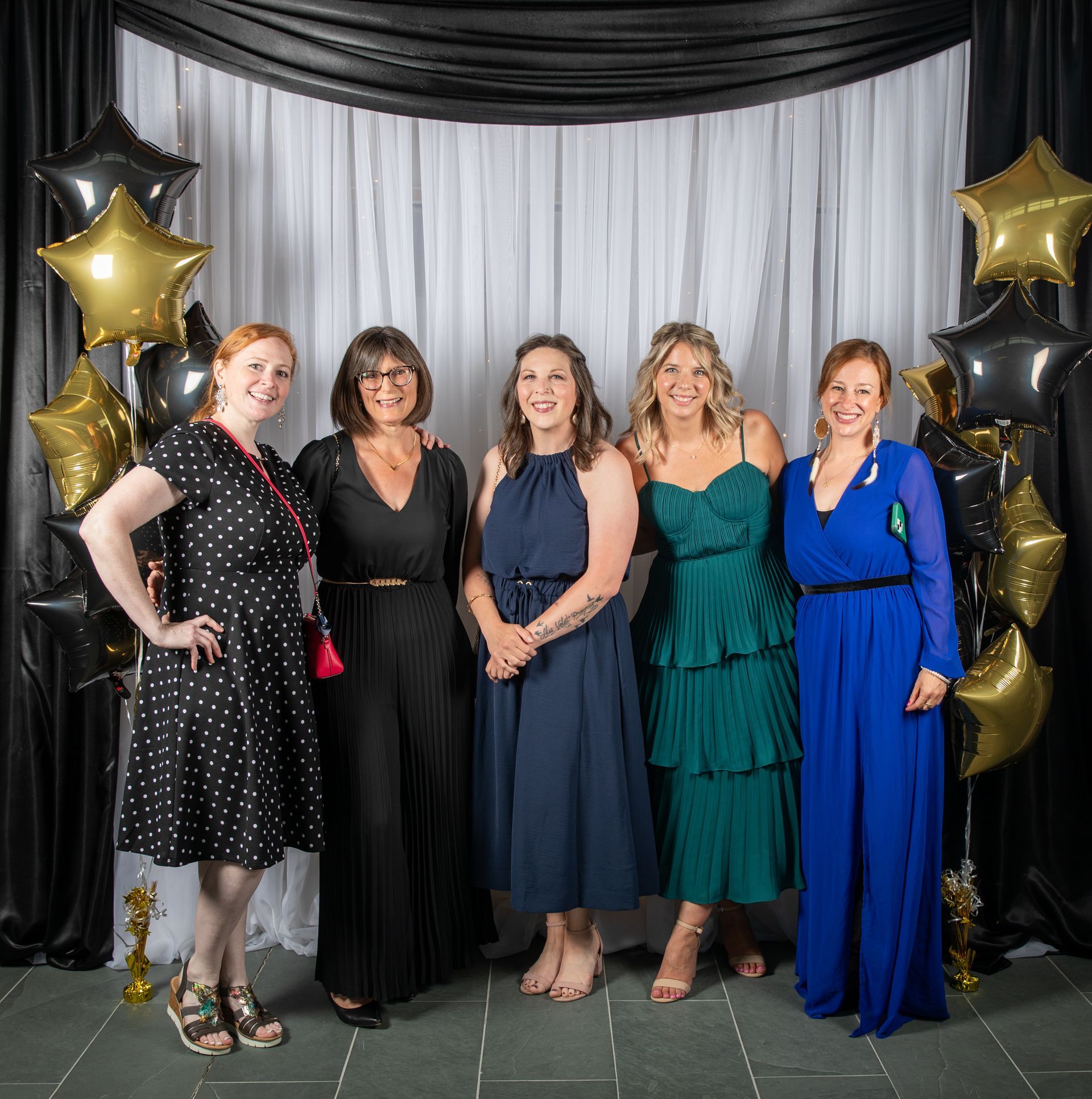 Five women in formal wear pose in front of a black and white backdrop with gold star balloons.
