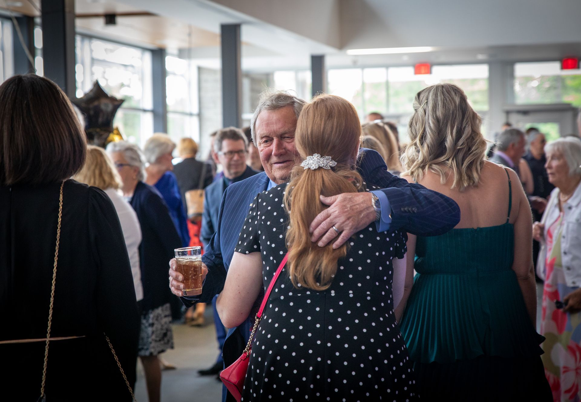 Man hugs woman at an event; he holds a drink. Other guests in the background.