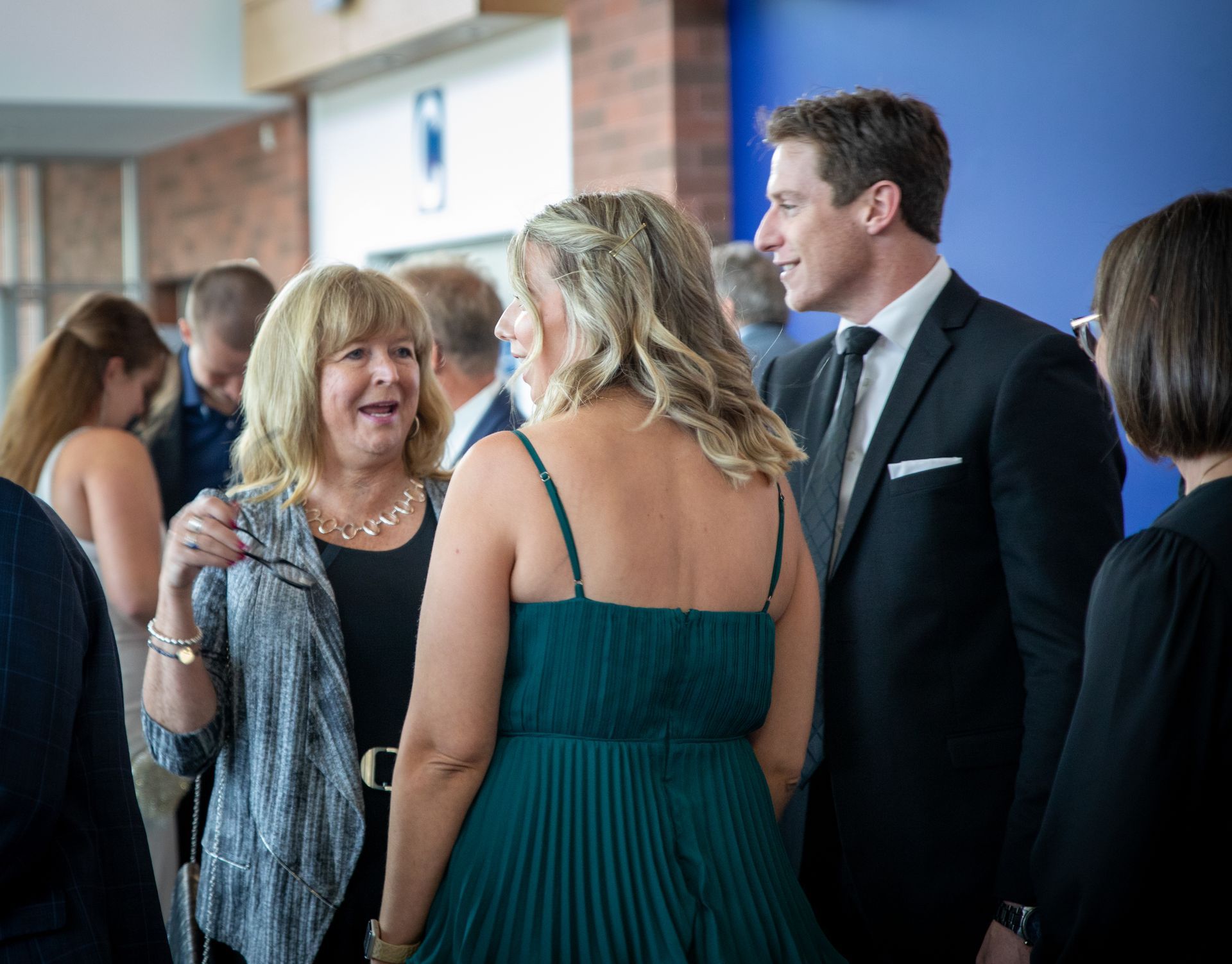 A group of people in a hall talking. A woman in a green dress is smiling, a man in a suit looks on.