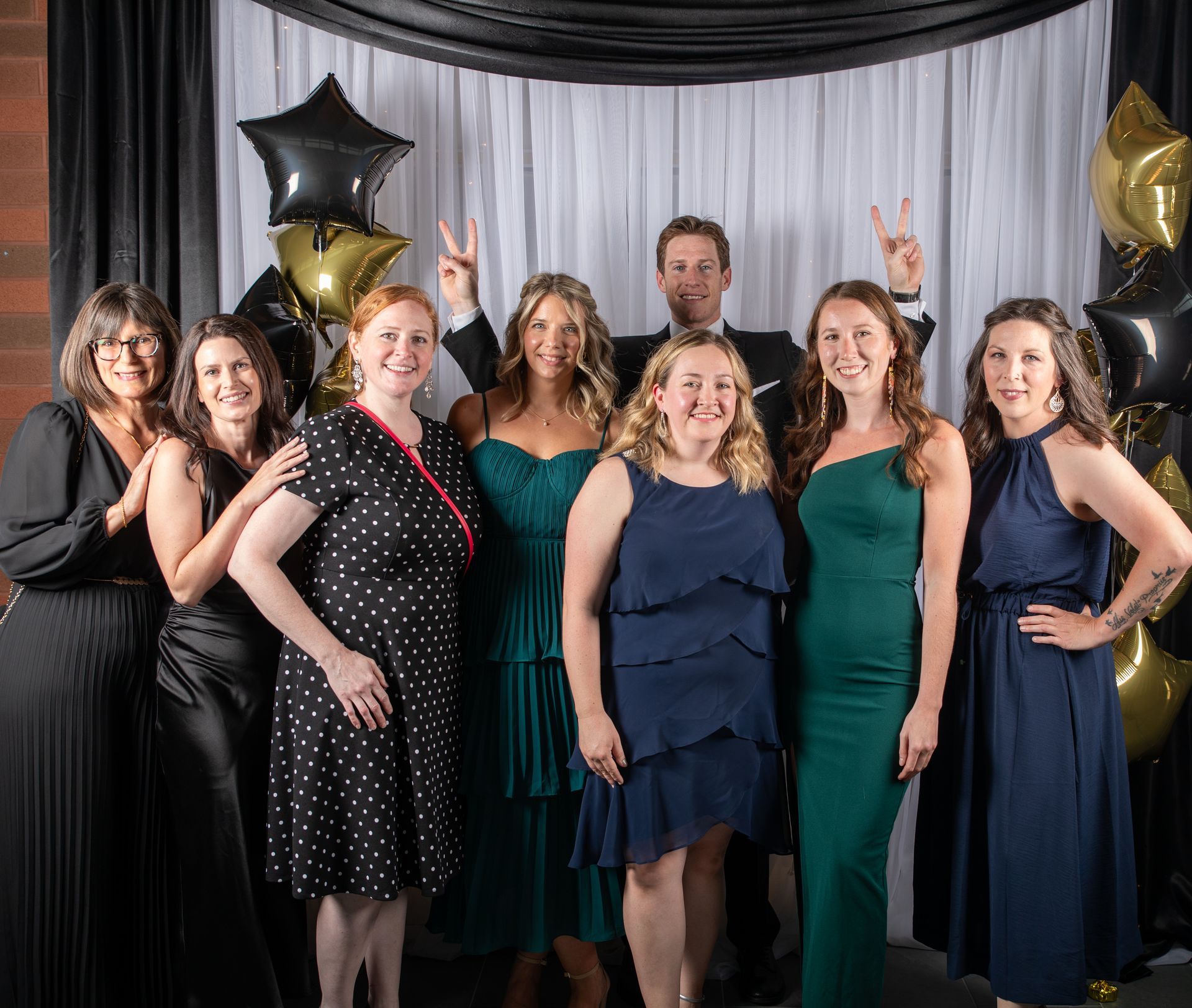 Group of people in formal attire pose for a photo. Some raise peace signs. Black and gold balloons in background.