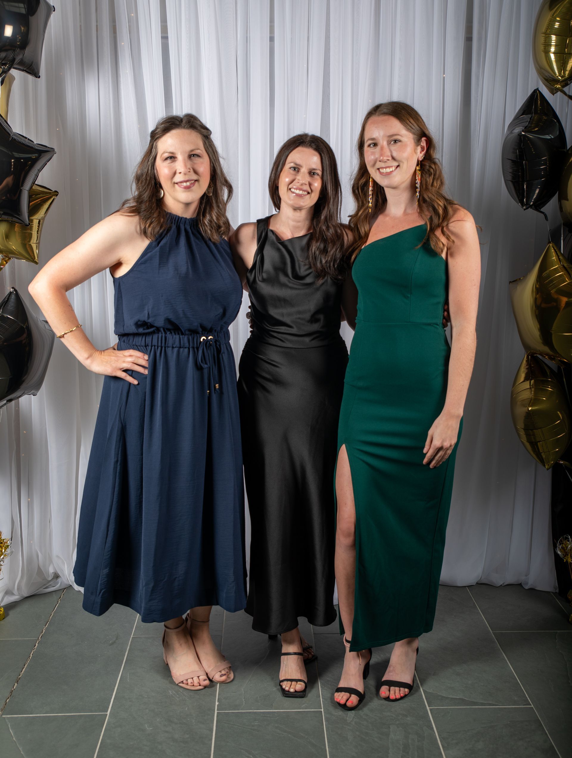 Three women in evening dresses posing together against a white backdrop with balloons.