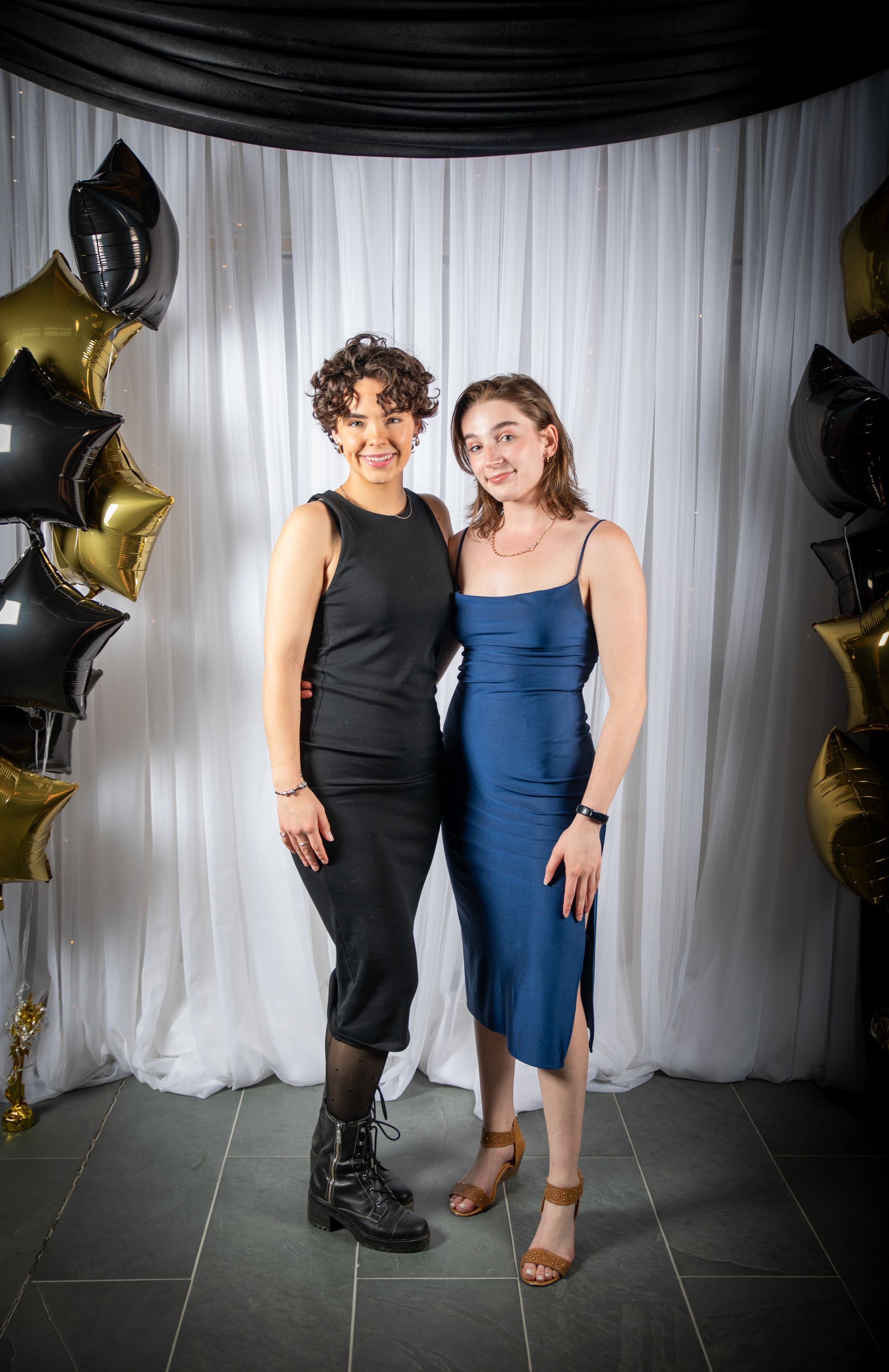 Two women pose in front of a white curtain backdrop with black and gold balloons.
