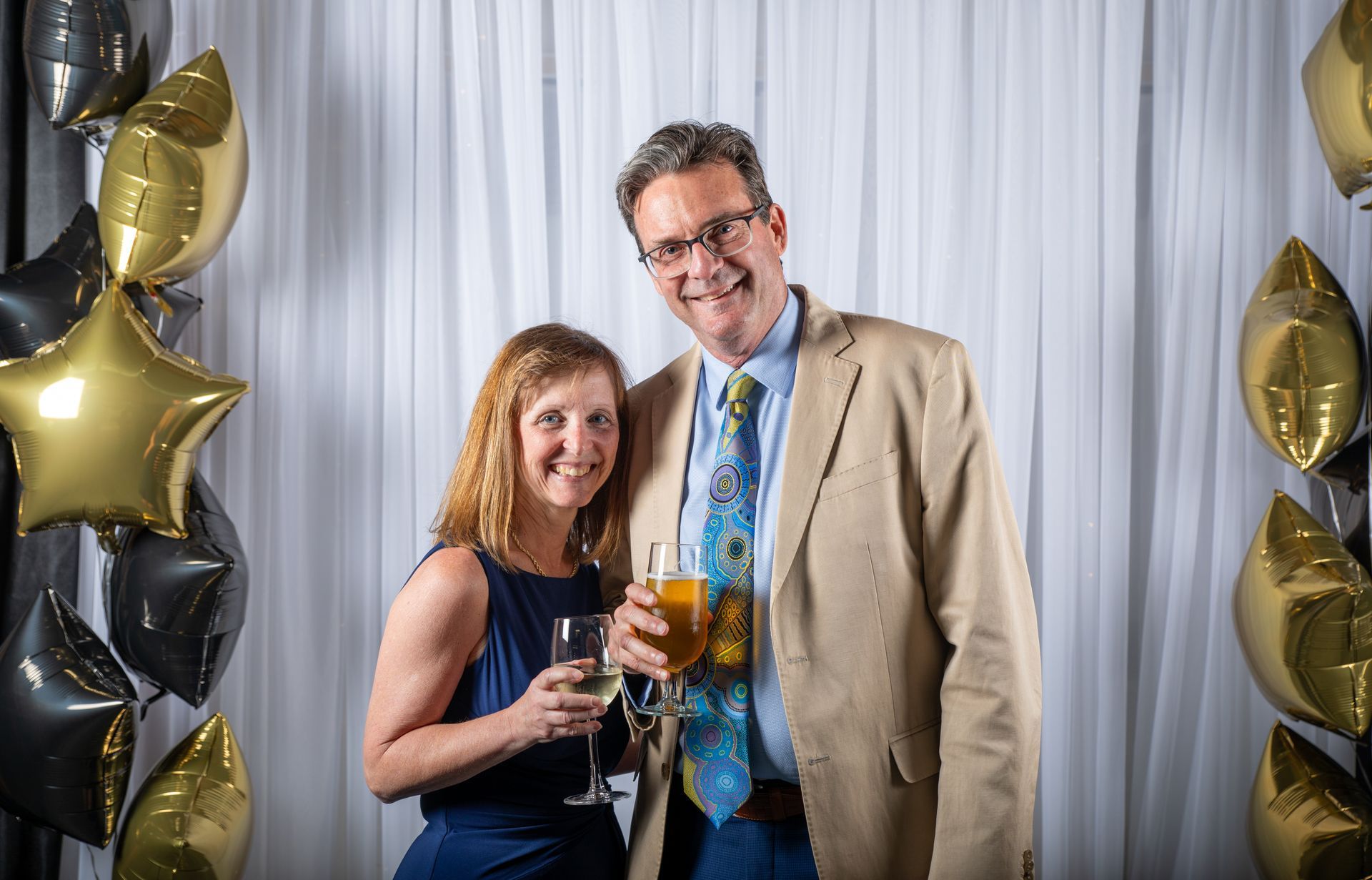 Couple smiling, holding drinks, posing in front of a curtain backdrop with gold and black balloons.
