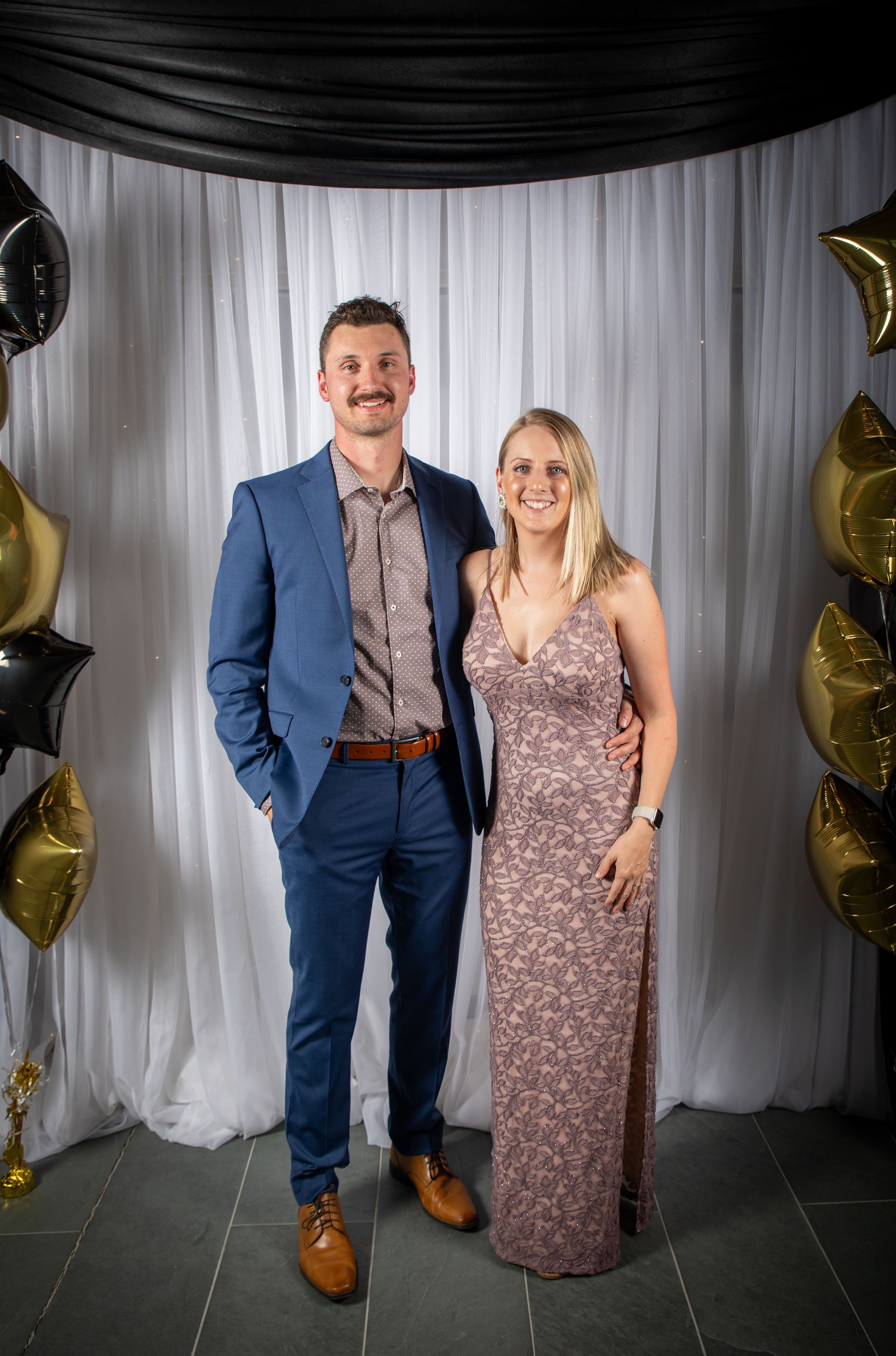 Man and woman posing formally. He in blue suit, she in pink gown. Standing by a draped backdrop with balloons.