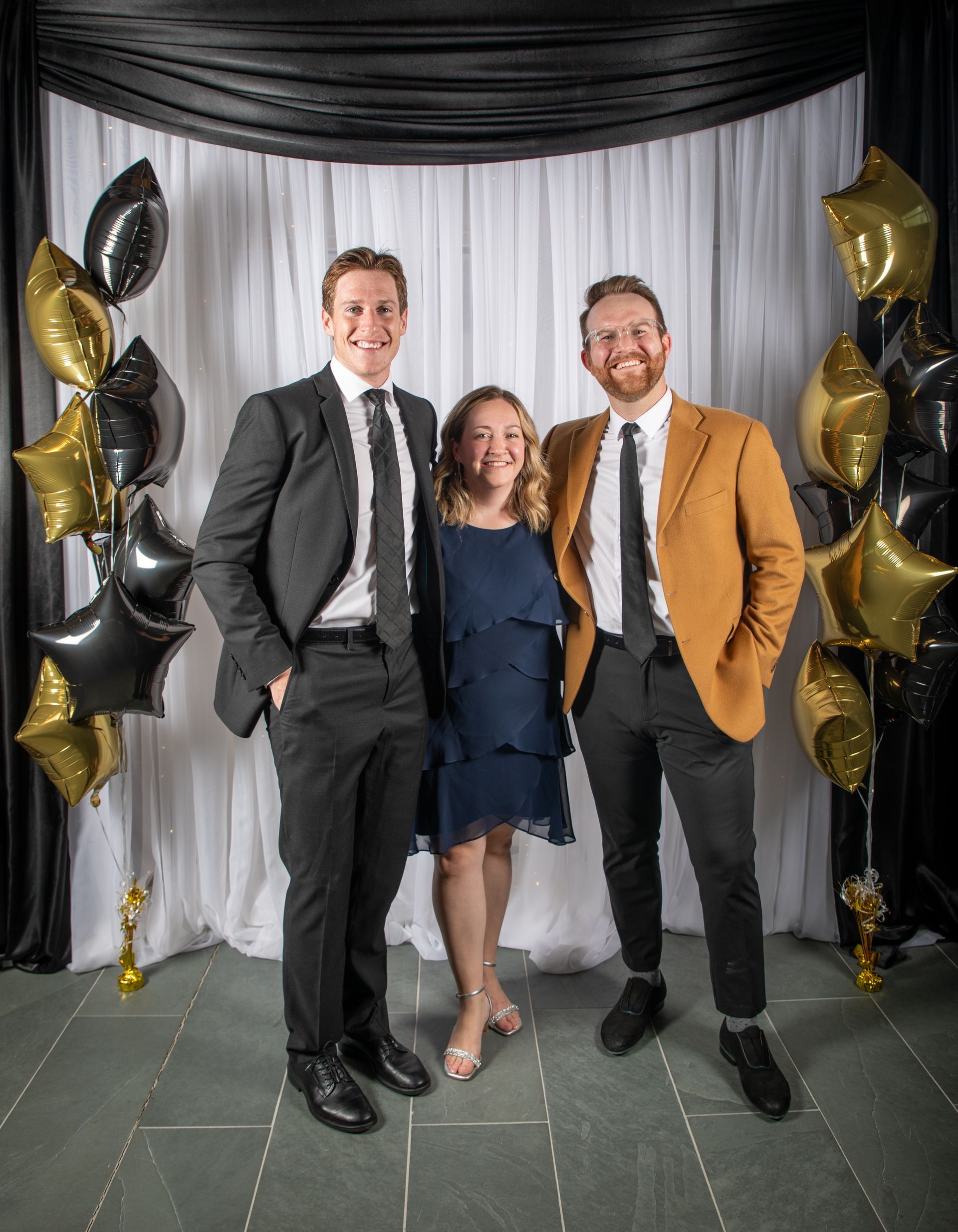 Three people pose in front of a backdrop with balloons; man in suit, woman in dress, man in blazer.